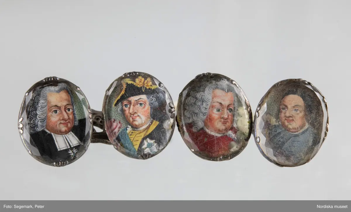 Manschettknappar, två par, miniatyrer. 1700-tals mitt.  Silver, glas.
Saknar stämplar. På varje knapp ett ovalt porträtt i miniatyr i silverinfattning, baksidan räfflad, knapparna förenade två och två, på den ena föreningslänken. ”I H” i genombrutit arbete, den andra föreningslänken senare ersättning. Manschettknapparna hölls i ett rött marokäng (getskinn) etui.
(katalogiseringen gäller a-c)