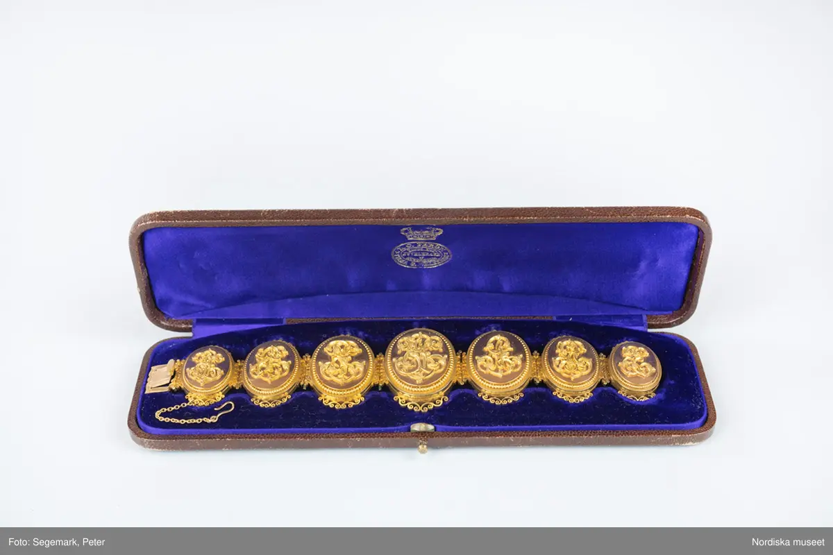 A) Armband, av matt och blankt guld, bestående av sju stående, sammanlänkade medaljonger som innesluter fotografiporträtt under glas. På varje medaljonglock ett pålagt monogram (för- och efternamnsinitialer) samt runt ytterkanten en smal pärlstav.  Genombruten dekor upptill och nedtill. Släta baksidor. Lås med bred snäppare stämplad med firmanamn, riks-, karat-, och stadsstämpel samt årsbokstav. Säkerhetskedja, med hake. Tillverkat i Stockholm 1875 av L C Féron/firma Gustaf Möllenborg. 

B) Originaletui, avlångt, välvt, med rundade hörn, klätt med brunt mönsterpressat skinn. Fällock med tryckknäppe av mässing. Etuiets insida klädd med lila sammet och siden (locket). Firmastämpel i guldtryck.

Porträtten föreställer Victor Edman (4) i centrum, omgiven av sina sex barn. På vänster sida finns de två äldsta barnen (2) och (3) från hans första äktenskap med Ulrika Charlotta Edman, f. Lagerhjelm (1818-1846). Längst ut på vänster sida finns det första barnet (1) i hans andra äktenskap med Emilia Elisabeth (Elise) Edman, f. Biel, en son som dog i unga år. På höger sida, de yngre barnen (5) (6) och (7), med Elise Edman:
1. VE: Johan Victor Edman, student, f. 12/8 1852, d. 6/3 1866.
2. CE: Carl Gustaf Edman, kanslisekreterare i civildepartementet, f. 26/7 1846, d. 3/5 1921. 
3. PE: Pehr Victor Edman, kapten vid Västmanlands regemente, f. 10/10 1841, d. 7/12 1879.
4. VE: Beställaren av armbandet, familjefadern Victor Edman, överstelöjtnant, f. 29/9 1813, d. 6/10 1880.  I locket en ljus lock av Victor Edmans hår. 
5. AE: Anna Mathilda Elisabeth Edman, f. 4/6 1856, d. 13/9 1882. I locket ett porträtt av maken, Anders Axel Altberg, godsägare (1839-1902). 
6. GE: Fredrik Gillis Edman, agronom, f. 6/7 1857, d. 19/11 1902.
7. EE: Hedvig Emilia (Emy) Edman, f. 2/5 1865, d. 11/9 1935, senare trägravör. Gift 1892 med Johan (John) Ludvig Kindborg, konstnär (1861-1907). 
/Anna Womack 2017-08-18