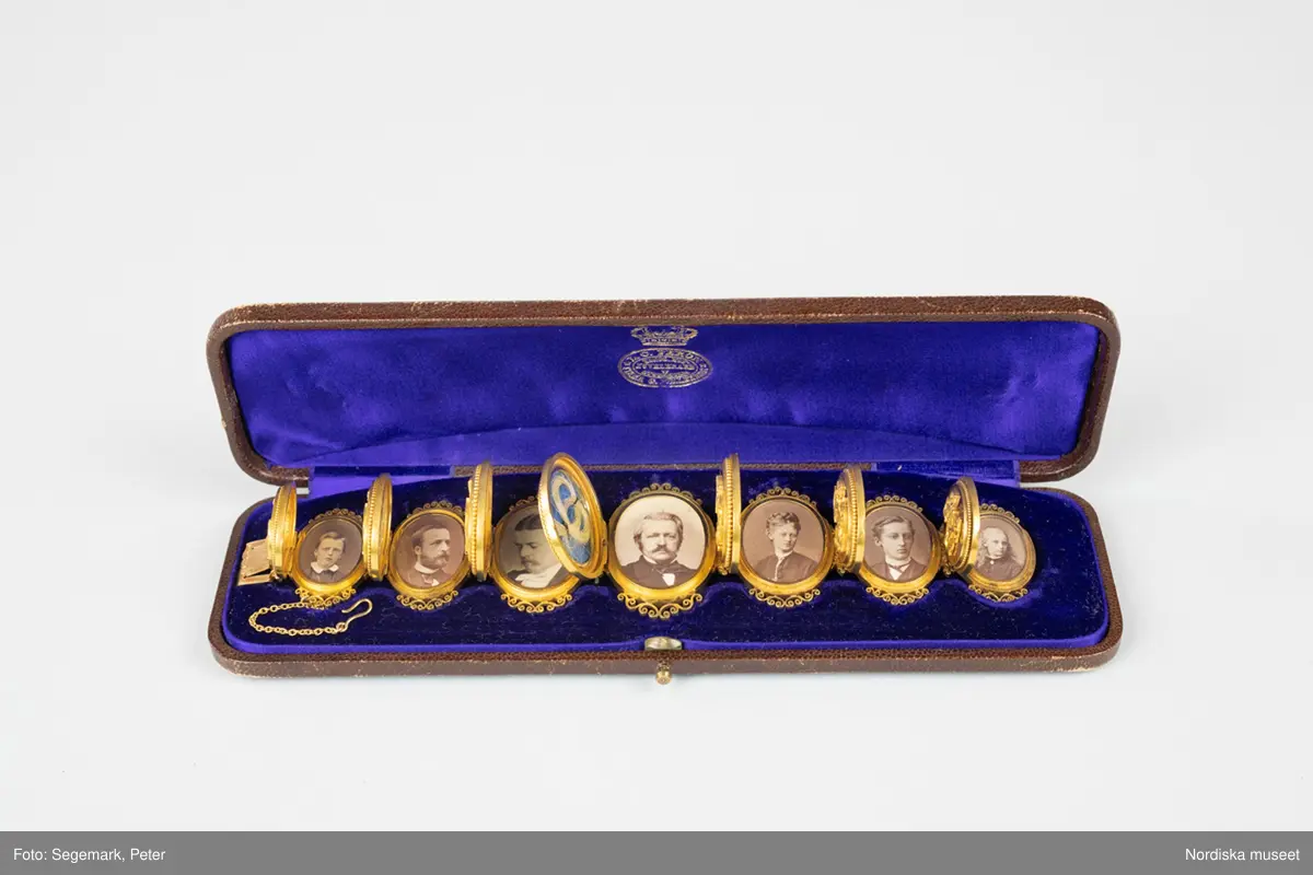 A) Armband, av matt och blankt guld, bestående av sju stående, sammanlänkade medaljonger som innesluter fotografiporträtt under glas. På varje medaljonglock ett pålagt monogram (för- och efternamnsinitialer) samt runt ytterkanten en smal pärlstav.  Genombruten dekor upptill och nedtill. Släta baksidor. Lås med bred snäppare stämplad med firmanamn, riks-, karat-, och stadsstämpel samt årsbokstav. Säkerhetskedja, med hake. Tillverkat i Stockholm 1875 av L C Féron/firma Gustaf Möllenborg. 

B) Originaletui, avlångt, välvt, med rundade hörn, klätt med brunt mönsterpressat skinn. Fällock med tryckknäppe av mässing. Etuiets insida klädd med lila sammet och siden (locket). Firmastämpel i guldtryck.

Porträtten föreställer Victor Edman (4) i centrum, omgiven av sina sex barn. På vänster sida finns de två äldsta barnen (2) och (3) från hans första äktenskap med Ulrika Charlotta Edman, f. Lagerhjelm (1818-1846). Längst ut på vänster sida finns det första barnet (1) i hans andra äktenskap med Emilia Elisabeth (Elise) Edman, f. Biel, en son som dog i unga år. På höger sida, de yngre barnen (5) (6) och (7), med Elise Edman:
1. VE: Johan Victor Edman, student, f. 12/8 1852, d. 6/3 1866.
2. CE: Carl Gustaf Edman, kanslisekreterare i civildepartementet, f. 26/7 1846, d. 3/5 1921. 
3. PE: Pehr Victor Edman, kapten vid Västmanlands regemente, f. 10/10 1841, d. 7/12 1879.
4. VE: Beställaren av armbandet, familjefadern Victor Edman, överstelöjtnant, f. 29/9 1813, d. 6/10 1880.  I locket en ljus lock av Victor Edmans hår. 
5. AE: Anna Mathilda Elisabeth Edman, f. 4/6 1856, d. 13/9 1882. I locket ett porträtt av maken, Anders Axel Altberg, godsägare (1839-1902). 
6. GE: Fredrik Gillis Edman, agronom, f. 6/7 1857, d. 19/11 1902.
7. EE: Hedvig Emilia (Emy) Edman, f. 2/5 1865, d. 11/9 1935, senare trägravör. Gift 1892 med Johan (John) Ludvig Kindborg, konstnär (1861-1907). 
/Anna Womack 2017-08-18