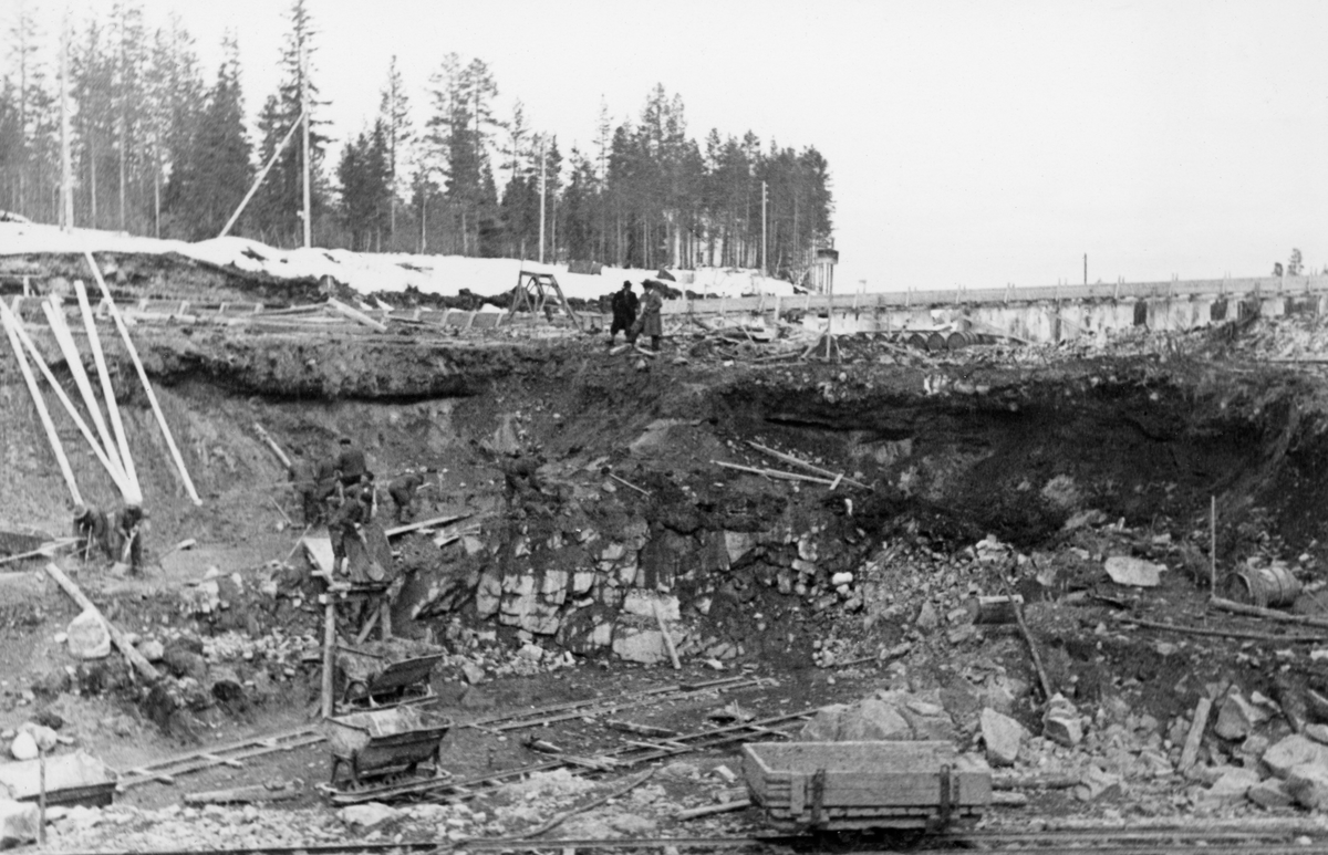 Fra anleggsområdet der det i perioden 1936-1938 ble reist en ny reguleringsdam ved elva Søndre Osas utløp fra Osensjøen i Åmot kommune i Hedmark. Dette fotografiet ble tatt tidlig i april 1937, antakelig der dammen skulle bygges. Anleggsarbeiderne hadde i hvert fall gravd seg djupt ned, gjennom et sand- og gruslag og ned i ei sone med sprukkent fjell. Da dette fotografiet ble tatt arbeidet åtte menn med spader og trillebårer på en avsats på nordsida av negdskjæringa. Ved ytterkanten av denne avsatsen var det oppsatt ei rampe av tre. Derfra kunne arbeiderne tippe trillebårene sine ned i ei tralle, ei skinnegående vogn, som, når den ble full, antakelig kunne skyves til en «tipp» lengre nede ved elvebredden. På kanten mot nedgravinga, sentralt i bildet sto to frakkekledde menn i samtale, med ansiktene vendt mot anleggsområdet og arbeiderne. Bak dem skimter vi to forskalinger, ei på tvers av anleggsområdets lengderetning og ei langsgående. Den sistnevnte kan ha vært ei slags omløpsrenne der vannet og tømmeret fra Osensjøen kunne passere uten at det forstyrret aktiviteten i anleggsområdet.