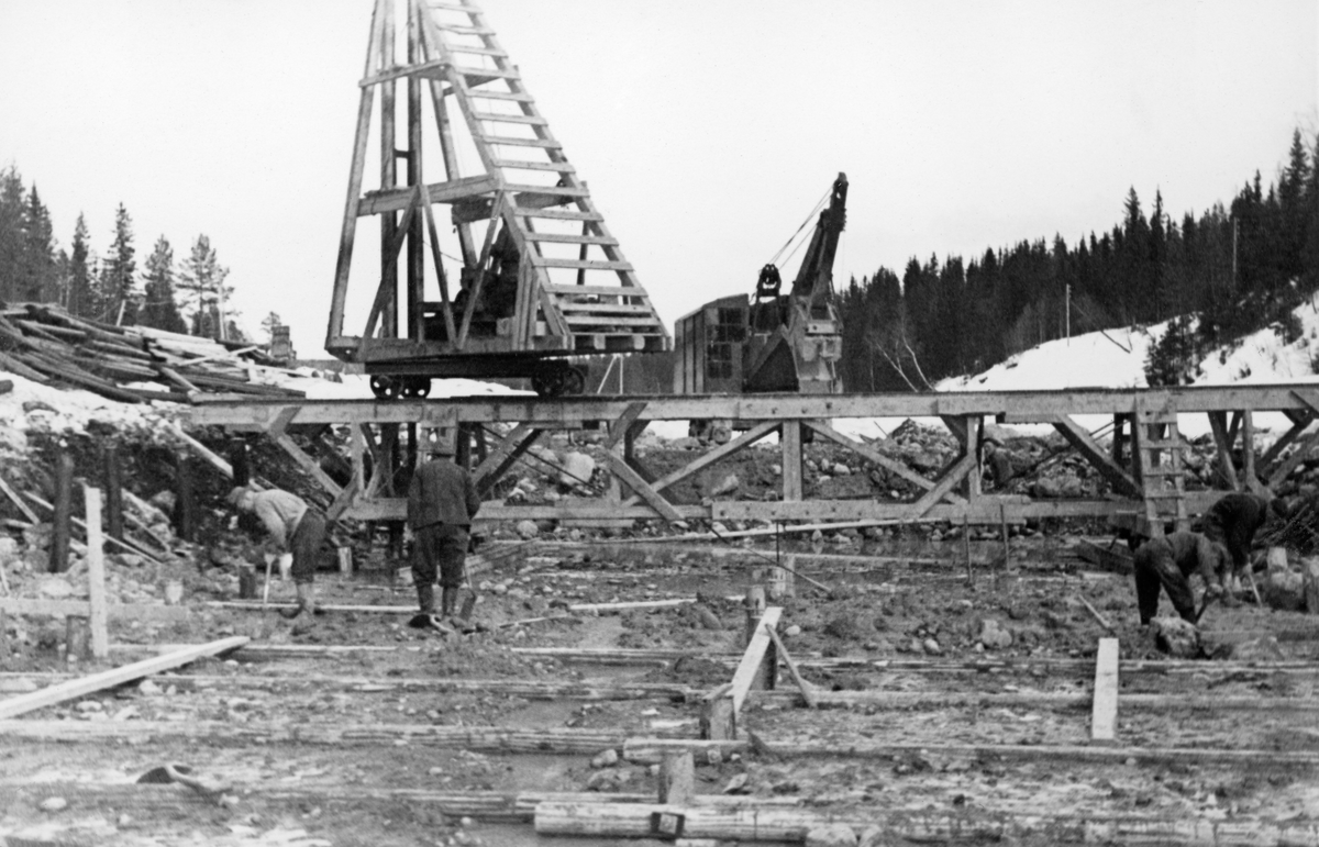 Fra bygginga av den nye dammen ved elva Søndre Osas utløp fra Osensjøen i Åmot kommune i Hedmark. Dette fotografiet er tatt tidlig i april i 1937, på et tidspunkt da det fortsatt lå snø i omkringliggende terreng. I forgrunnen arbeidet man antakelig med det som skulle bli et nytt løp for Søndre Osa ved det nye damstedet. I forgrunnen arbeidet fire karer med spader på et areal som antakelig skulle planeres. På marka nærmest fotgrafen var det slått ned en del pæler som det var fastskrudd stolper til, med regelmessig innbyrdes avstand. Umiddelbart bak anleggsarbeiderne var det lagt ei provisorisk bru på tvers av anleggsområdet. På den sto det ei skinnegående tralle med et påbygd tretårn, sannsynlig vis en slags pælebukk. I bakgrunnen skimter vi en gravemaskin, som antakelig tok unna de groveste massene, før karene med spadene gjorde «finishen».