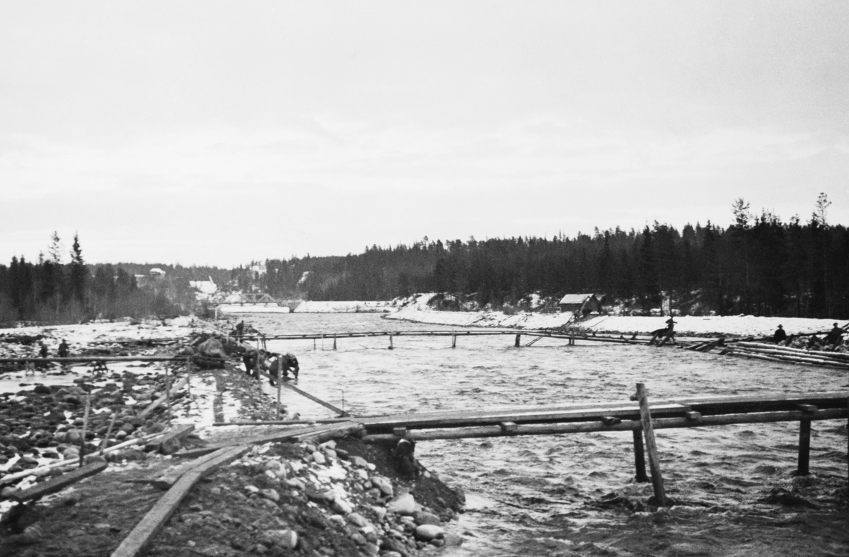 Den nedre delen av elva Søndre Osa i Åmot kommune, fotografert i desember 1935, på et tidspunkt da man arbeidet med å bygge voller langs begge sider av elveløpet. Her rant elva over en slaktskrånende mo der det rennende vannet, blant annet i forbindelse med dambruddet ved Osa-kraftverket i 1916, hadde skylt med seg mye av jordsmonnet og etterlatt seg ett bredt, steinete far som antakelig var forholdsvis grunt, sjøl i flomperiodene. Dette må ha vanskeliggjort fløtinga. Det anleggsarbeidet vi ser på dette fotografiet dreide seg antakelig om å samle vannet i et smalere og djupere løp ved å flytte masse fra den foretrukne traseen ut på kantene. Arbeiderne hadde bygd et par provisoriske trebruer, slik at det skulle være enkelt å komme seg over elveløpet med ei trillebår. Et stykke lengre oppe skimter vi ei fagverksbru av stål, der Desetvegen over Osmoen krysset Søndre Osa.