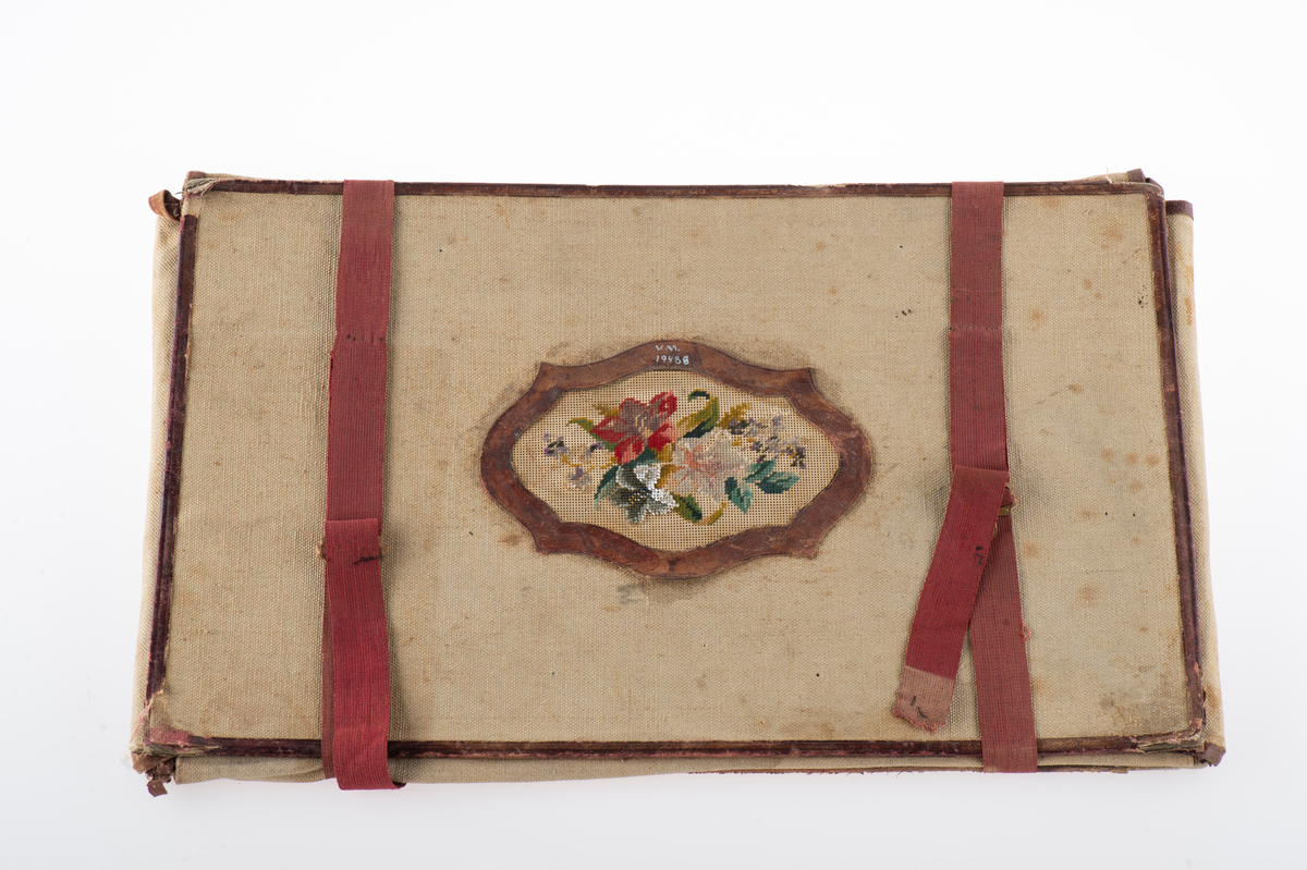 Av groft linne, kantat med brunt skinn, rödbruna bomullsband med spännen att binda det samman. I centrum en broderad utsmyckning föreställande blommor och blad.