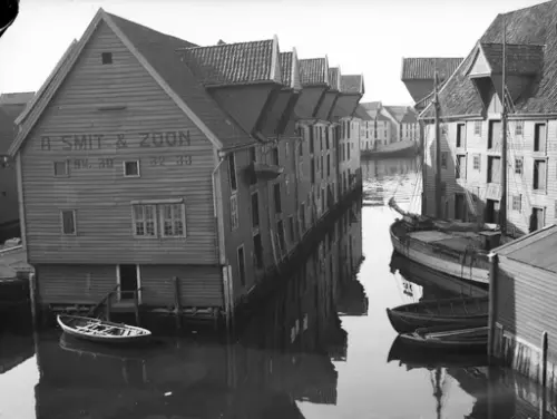 Sort-hvitt bilde av sjøboder i sjøen, og båter liggende i vannet like ved. 