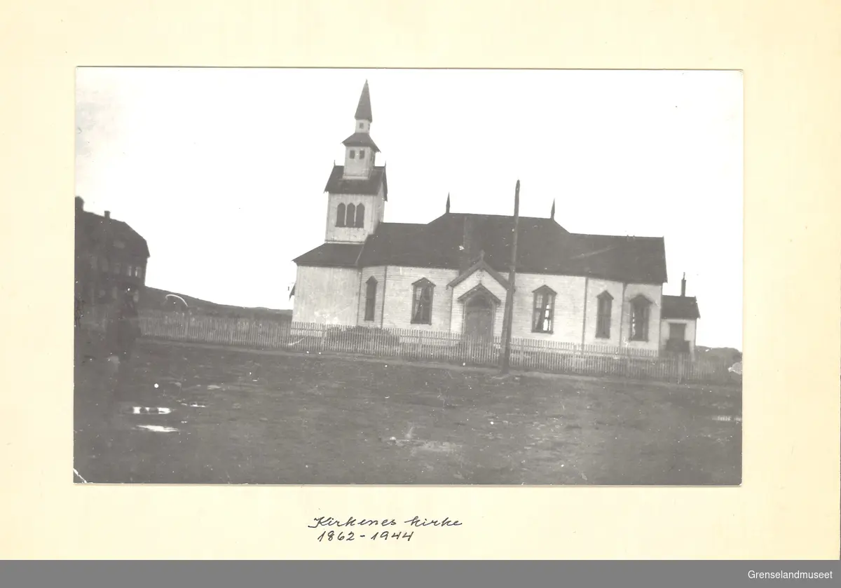 Svart-hvitt bilde av Kirken i Kirkenes, tatt mellom 1862 og 1944. Foto er tatt på skrått slikt at vinklingen blir skyvet på skrå mot høyre. Kirkenes kirke med tårn på venstre side av inngangen. Gjerdet rundt kirkegården og et avbildet hus til venstre, med knapt synlig person foran. 