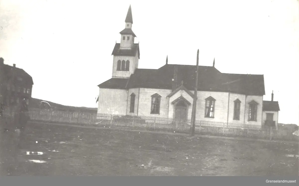 Svart-hvitt bilde av Kirken i Kirkenes, tatt mellom 1862 og 1944. Foto er tatt på skrått slikt at vinklingen blir skyvet på skrå mot høyre. Kirkenes kirke med tårn på venstre side av inngangen. Gjerdet rundt kirkegården og et avbildet hus til venstre, med knapt synlig person foran. 