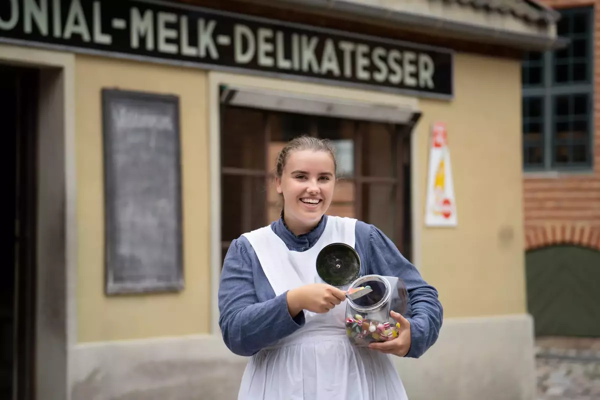 Smilende kvinne i historisk drakt står foran en kolonialbutikk på Norsk Folkemuseum. Hun holder krukke med gammeldagse drops og en metalløse, klar til å servere godteri.