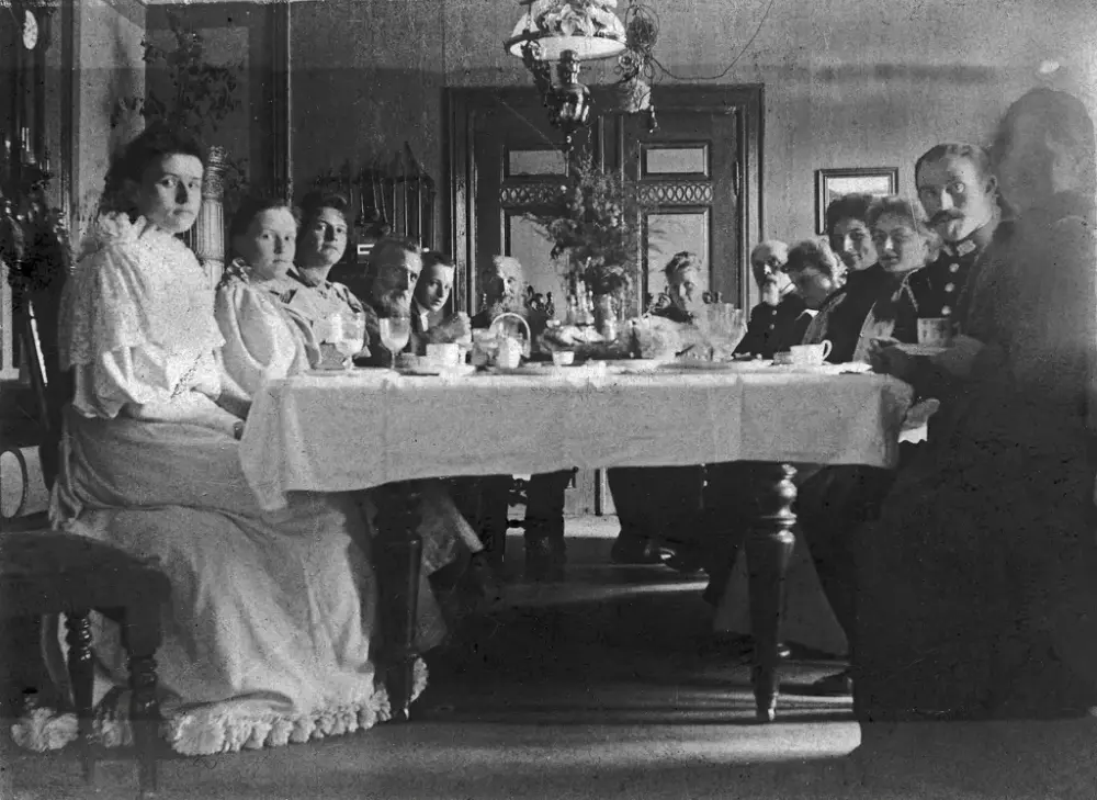 Svart hvitt foto fra  ca 1900 - spisestua på Gjøvik gård. Mange mennesker, pent kledde, sitter rundt det store spisebordet med kopper og servise foran seg, og ser mot fotografen.