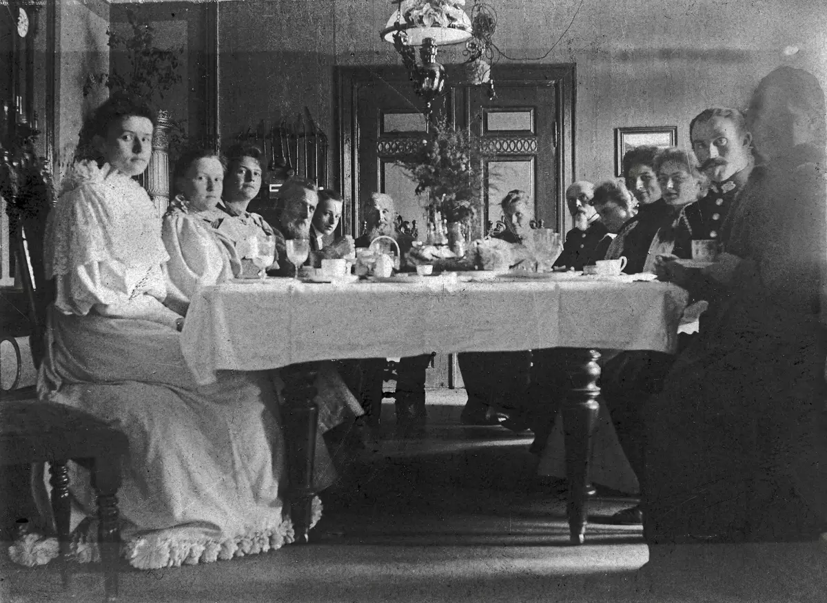 Svart hvitt foto fra  ca 1900 - spisestua på Gjøvik gård. Mange mennesker, pent kledde, sitter rundt det store spisebordet med kopper og servise foran seg, og ser mot fotografen.