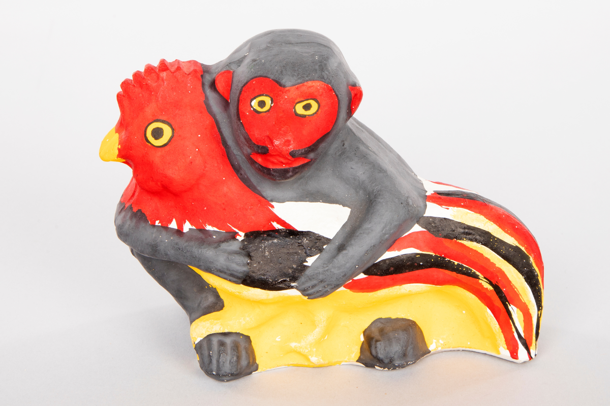 Figur i leire som forestiller en ape som holder rundt en hane. Apen er malt mørk grå med rødt ansikt og ører og gule øyne. Hanen er hvit og gul med rødt hode og kam, gult nebb og gule øyne. Stjert i rødt, svart, gult og hvitt.