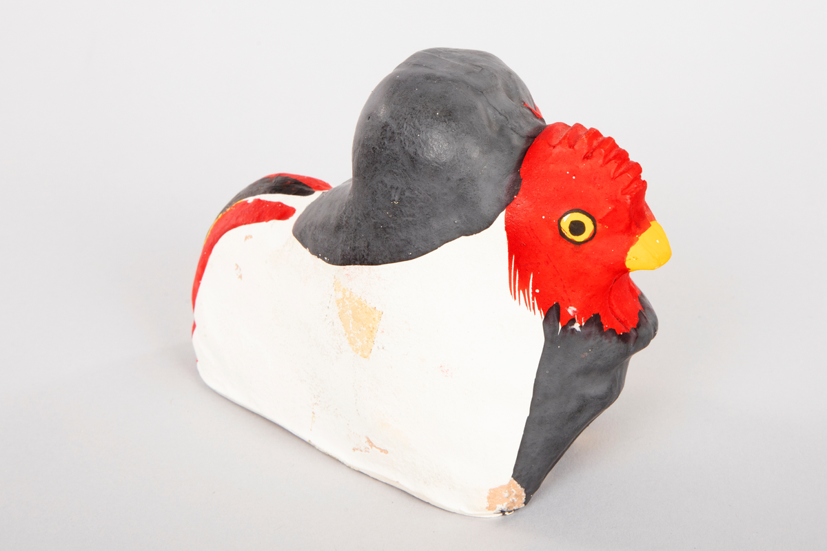 Figur i leire som forestiller en ape som holder rundt en hane. Apen er malt mørk grå med rødt ansikt og ører og gule øyne. Hanen er hvit og gul med rødt hode og kam, gult nebb og gule øyne. Stjert i rødt, svart, gult og hvitt.