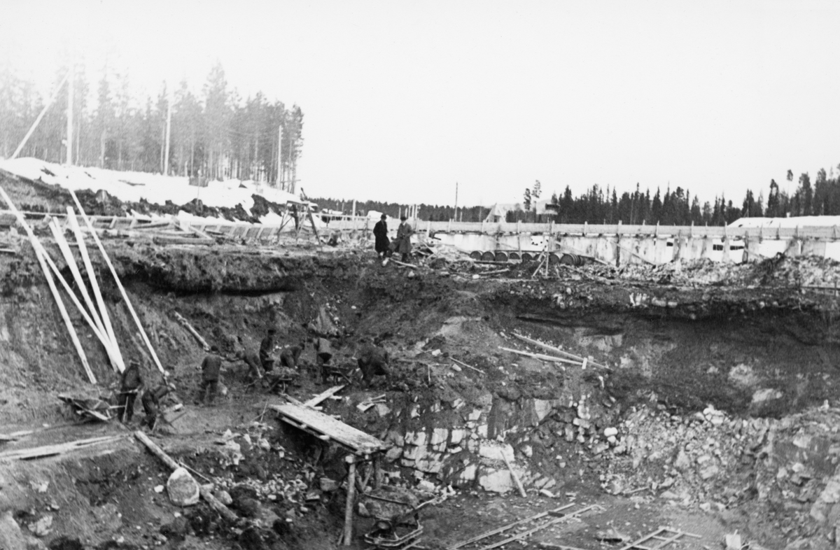 Fra anleggsområdet der det i perioden 1936-1938 ble reist en ny reguleringsdam ved elva Søndre Osas utløp fra Osensjøen i Åmot kommune i Hedmark. Dette fotografiet ble tatt tidlig i april 1937, antakelig der dammen skulle bygges. Anleggsarbeiderne hadde i hvert fall gravd seg djupt ned, gjennom et sand- og gruslag og ned i ei sone med sprukkent fjell. Da dette fotografiet ble tatt arbeidet åtte menn med spader og trillebårer på en avsats på nordsida av negdskjæringa. Ved ytterkanten av denne avsatsen var det oppsatt ei rampe av tre. Derfra kunne arbeiderne tippe trillebårene sine ned i ei tralle, ei skinnegående vogn, som, når den ble full, antakelig kunne skyves til en «tipp» lengre nede ved elvebredden. På kanten mot nedgravinga, sentralt i bildet sto to frakkekledde menn i samtale, med ansiktene vendt mot anleggsområdet og arbeiderne. Bak dem skimter vi to forskalinger, ei på tvers av anleggsområdets lengderetning og ei langsgående. Den sistnevnte kan ha vært ei slags omløpsrenne der vannet og tømmeret fra Osensjøen kunne passere uten at det forstyrret aktiviteten i anleggsområdet. Anleggsarbeidet sysselsatte fra 30-40 til 100 mann. Aktiviteten var størst når vannføringa i Søndre Osa var på det laveste. I den perioden da det var vårflom og fløting - i mai og juni - ble anleggsarbeidet langt på veg stanset.