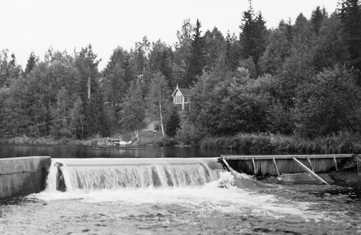 Strømsdammen i Nitelva i Nittedal kommune i Akershus, fotografert i motstrøms retning på seinsommeren i 1934, lenge etter at fløtingssesongen var over. Dette ser ut til å ha vært en terskeldam, altså en konstruksjon som først og fremst skulle løfte vannspeilet i den ovenforliggende delen av elva, for dermed å hindre at tømmeret satte seg fast der. Strømsdammen ser ut til å ha vært en forholdsvis lav betongmur med overløp noenlunde midt i elveleiet. Her var det cirka en meters fall. Damarmen til høyre i bildet var antakelig noe ustabil da fotografiet ble tatt, for der var damkrona forsterket med en trekonstruksjon på motstrøms side. Langs elvebreddene vokste det blandingsskog. I denne skogen skimter vi en glassveranda, som antakelig var en del av ei hytte. Fra hytta gikk det trapp ned til elvebredden, der det lå en liten, kvitmalt båt.