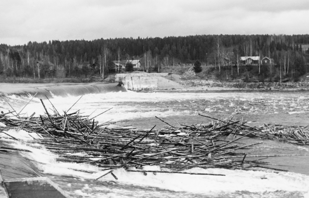 Kraftverksdammen ved Skjefstadfossen i Heradsbygda i Elverum.  Fotografiet er tatt fra vest på et tidspunkt da elva var flomstor, antakelig en vårdag i 1940-åra.  I forgrunnen ser vi at det har bygd seg opp en tømmervase (haug) på golvet under damkrona og på den nedenforliggende steinøra.  Lengre øst ser vi hvordan vannmassene i den djupeste delen av elveløpet strømmer over damkrona og ned i et underliggende basseng med kvitskummende overflate før det føres videre nedover et relativt bredt, urolig elveløp.  Der dammen møtte elvebredden på østsida var det murt og støpt et kraftig landkar med utsikt over dammen.  På sletta bak elvebrinken skimter vi bygningene på gardsbrukene Butteberg og Gropa.  I bakgrunnen lå det en skogkledd ås. 