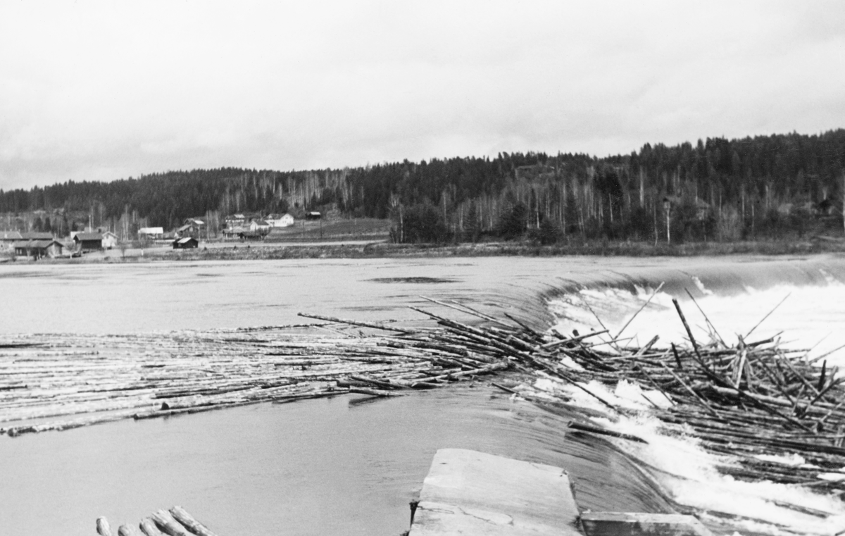 Tømmervaser på kanten av kraftverksdammen ved Skjefstadfossen i Heradsbygda i Elverum.  Fotografiet er tatt fra den vestre sida av anlegget, ikke langt fra inntaket til turbinrørene mot det nedenforliggende kraftverket.  Fotografiet viser en del tømmer som flyter mot damkanten, noe som har hengt seg på denne kanten og en haug som er blitt liggende på det skrå golvet nedenfor damfallet.  På motsatt side av elva, til venstre i bildet, ser vi gardsbruket Fossum. 