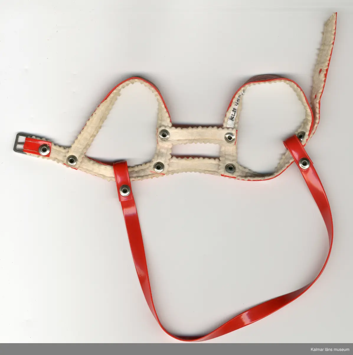 KLM 45519:16:3. Docksele, röd, av plast, konstfiber och metall. Plastband, 5 stycken, ihopnitade, 2 öglor för axlar och 2 band vid bröstkorg, ett kort och ett längre som spänns och fästs i ryggen med hål och skärpspänne. Plastögla, baktill, att hålla i. Bandet runt dockkroppen, fodrat. Dockan hade selen på sig vid lämnandet till KLM.