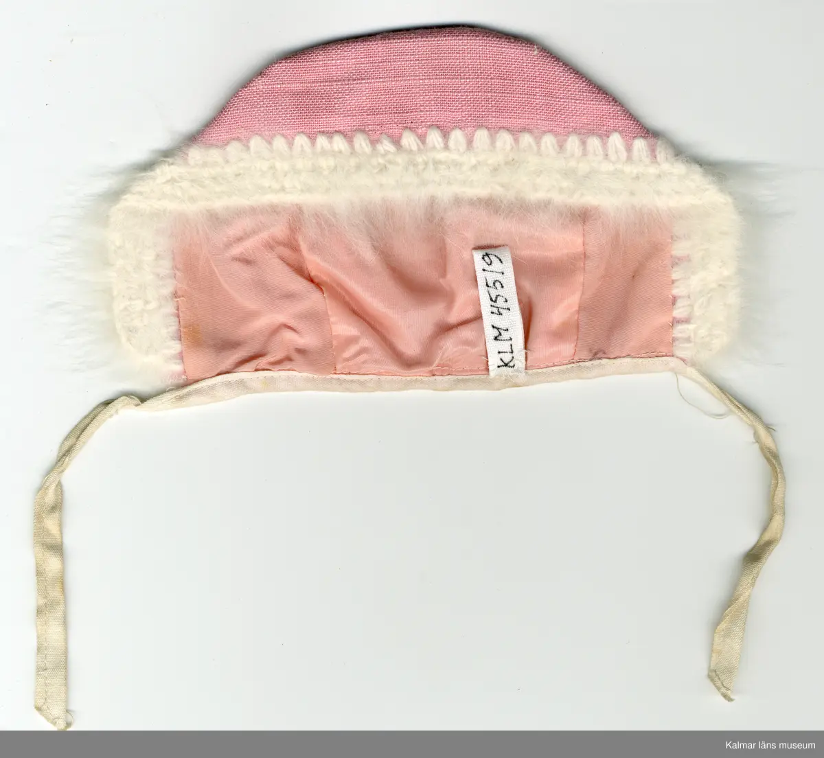 KLM 45519:16:2:1-2. Dockdräkt, overall med huvudbonad, av konstmaterial, bomull, angoragarn och metall, rosa och vit. :1 Overall, rosa, runt halslinning vitt kantband och vit dragkedja framtill. :2 Mössa, hätta, rosa, fodrad, kant, framtill, virkad med vitt angoragarn, kant, baktill, sydd med vitt kantband, som framtill blir knytband. Dockan hade båda plaggen på sig vid lämnandet till KLM.