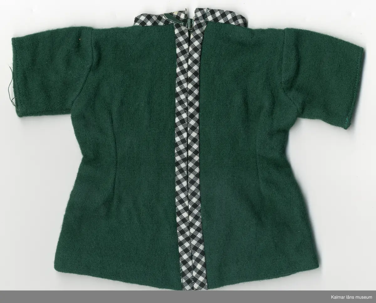 KLM 45519:17:5. Dockklänning, av textil, ylle och bomull, grön. Ärm, kort, midja, insydd både framtill och baktill. Halslinning kantad med bomullstyg, svart och vitrutigt, sytt som kantband, som bildar en rund kragkant vid hals, samma kantband fortsätter lodrätt ner över ryggstycket, på var sida om dragkedjan, vilken döljs. Givaren, Ann-Sofie Karlsson, bar en likadan klänning som barn.