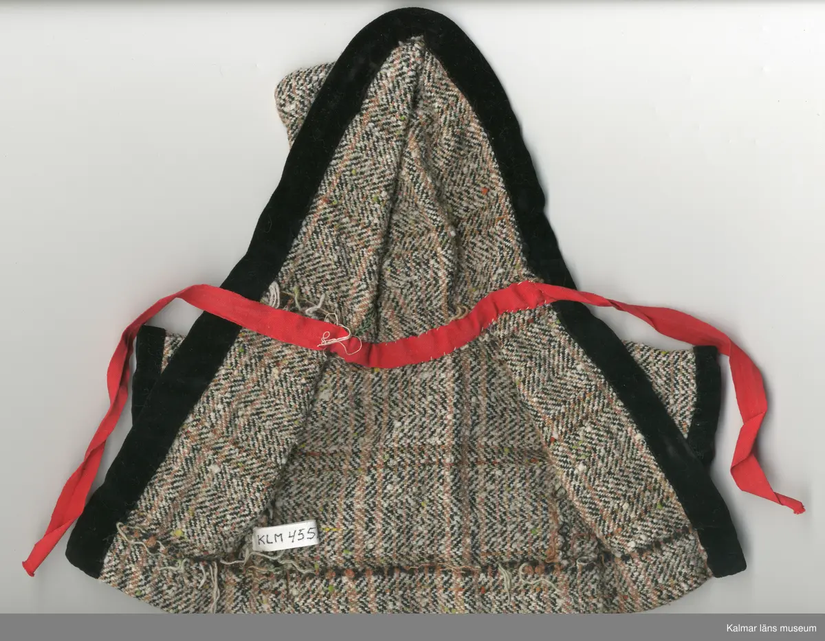 KLM 45519:16:10. Dockkappa, med huva, av konstmaterial, brun, vit och svartmelerad. Kappa kantad, framtill, även huva och ärm, nertill, med sammetskant, svart. Under huva, inuti kappa, bomullsband, rött, fastsytt med lösa ändar, som knyts