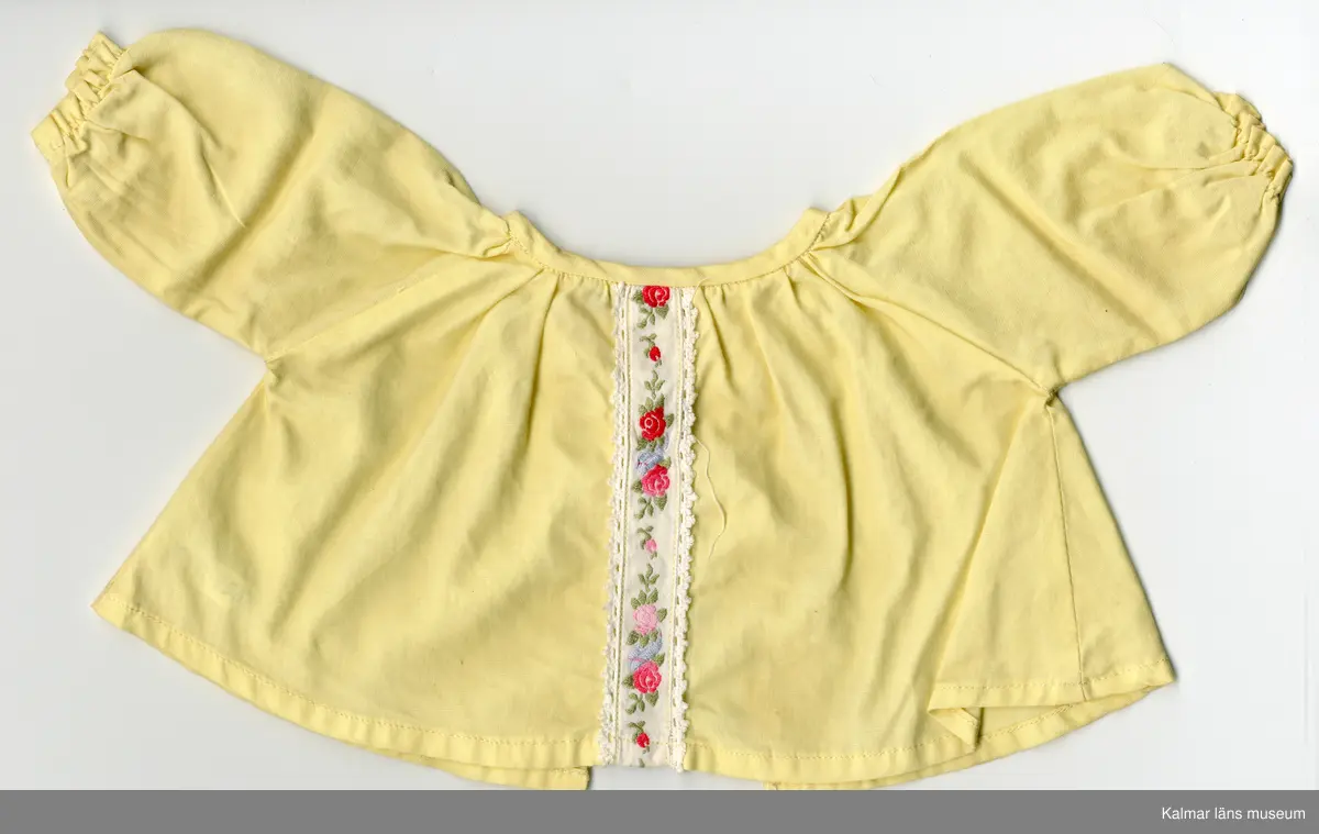 KLM 45519:17:7:1-2. Dockdräkt, :1 Klänning och : 2 Underbyxor, båda plaggen av textil, bomull, gul. :1 Klänningens ärm, lång, rynkad, resårband nertill. Klänning, rynkad, krage kantad med gult kantband, i samma tyg som klänningen, som knäpps i nacke med tryckknapp. Framtill, från krage till fåll, vitt band med spetskant och rosenbård i rött, grönt, blått och rosa. :2 Underbyxa med korta ben, rynkad, resår i midja och nertill vid ben. Lappar i vitt band med röd siffra, sydda inuti plaggen, antyder att plaggen är fabriksgjorda.