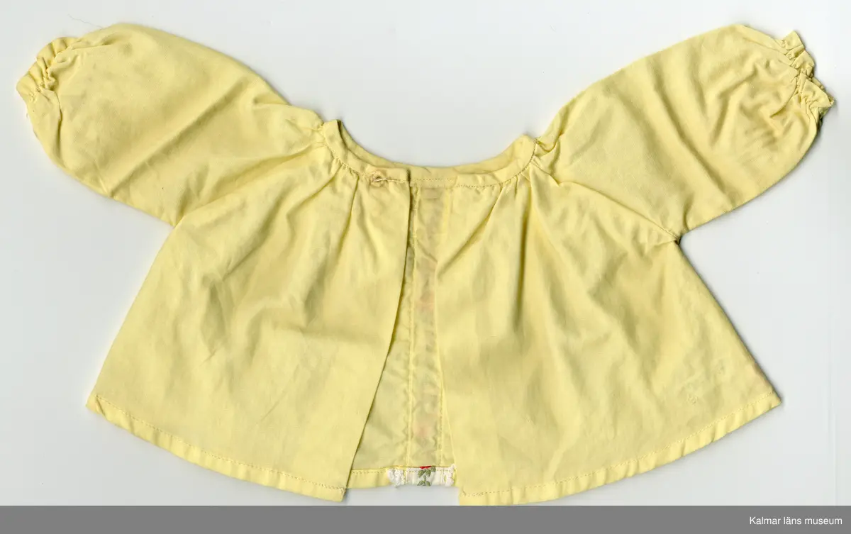 KLM 45519:17:7:1-2. Dockdräkt, :1 Klänning och : 2 Underbyxor, båda plaggen av textil, bomull, gul. :1 Klänningens ärm, lång, rynkad, resårband nertill. Klänning, rynkad, krage kantad med gult kantband, i samma tyg som klänningen, som knäpps i nacke med tryckknapp. Framtill, från krage till fåll, vitt band med spetskant och rosenbård i rött, grönt, blått och rosa. :2 Underbyxa med korta ben, rynkad, resår i midja och nertill vid ben. Lappar i vitt band med röd siffra, sydda inuti plaggen, antyder att plaggen är fabriksgjorda.