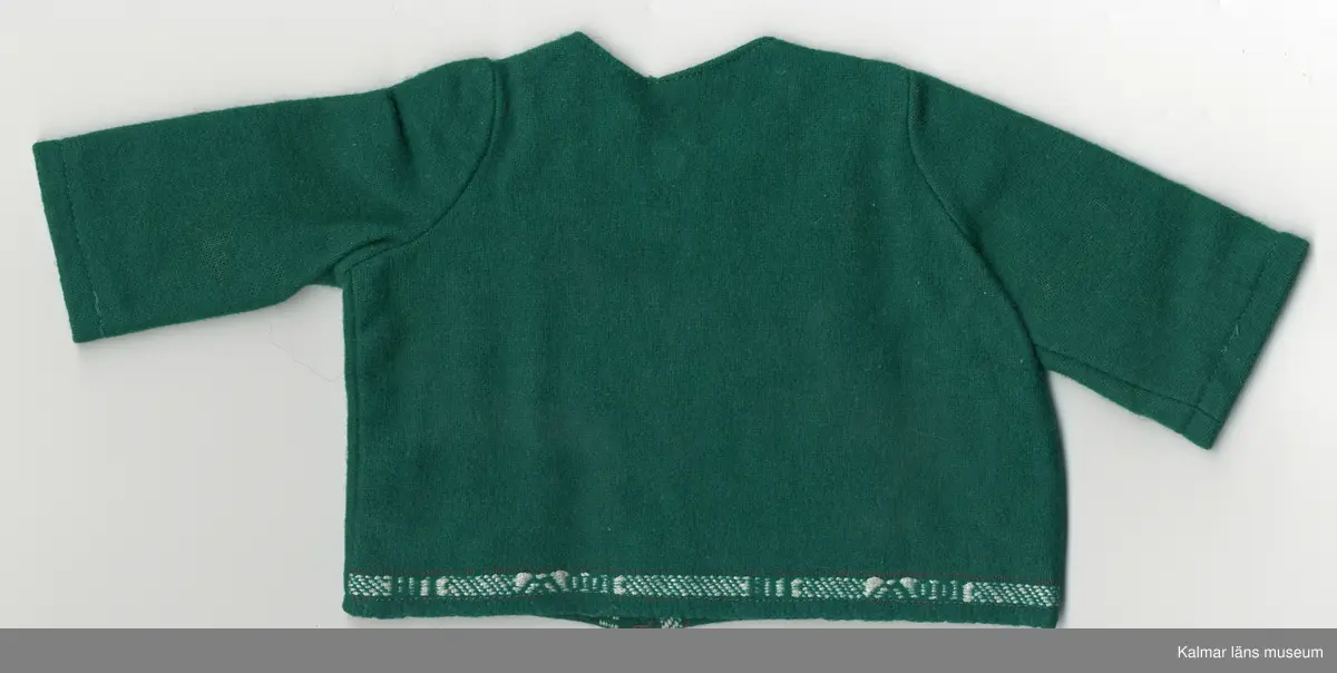 KLM 45519:17:10: 1-3. Dockdräkt, 3 plagg av textil, ylle, grönt. Mönsterbård, invävd, vit och grön med text: ALL WOOL. :1 Dockjacka, rundad halsurringning. dragkedja, framtill, vit. Mönsterbård, två stycken, framtill, längs dragkedja och en bak på jacka, nere vid fållen. :2 Dockbyxa, "sparkbyxa", byxa, hopsydd med rundning vid tår, vänster ben längre. Midja, linning med mönsterbård, knäpps i midja med hake och hyska, av metall. :3 Dockhuvudbonad, skärmmössa, av grönt ylle. Kulle och kant invändigt, förstärkt med Vlieseline, vitt. Mössa, kant, dekorerad med mönsterbård.