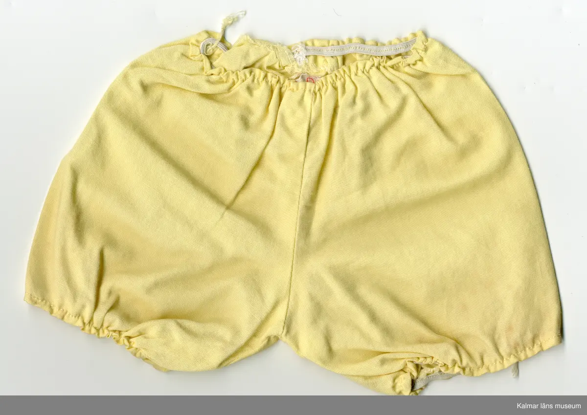 KLM 45519:17:7:1-2. Dockdräkt, :1 Klänning och : 2 Underbyxor, båda plaggen av textil, bomull, gul. :1 Klänningens ärm, lång, rynkad, resårband nertill. Klänning, rynkad, krage kantad med gult kantband, i samma tyg som klänningen, som knäpps i nacke med tryckknapp. Framtill, från krage till fåll, vitt band med spetskant och rosenbård i rött, grönt, blått och rosa. :2 Underbyxa med korta ben, rynkad, resår i midja och nertill vid ben. Lappar i vitt band med röd siffra, sydda inuti plaggen, antyder att plaggen är fabriksgjorda.
