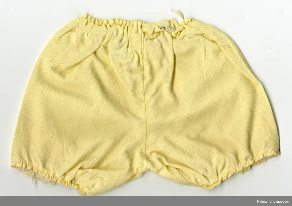 KLM 45519:17:7:1-2. Dockdräkt, :1 Klänning och : 2 Underbyxor, båda plaggen av textil, bomull, gul. :1 Klänningens ärm, lång, rynkad, resårband nertill. Klänning, rynkad, krage kantad med gult kantband, i samma tyg som klänningen, som knäpps i nacke med tryckknapp. Framtill, från krage till fåll, vitt band med spetskant och rosenbård i rött, grönt, blått och rosa. :2 Underbyxa med korta ben, rynkad, resår i midja och nertill vid ben. Lappar i vitt band med röd siffra, sydda inuti plaggen, antyder att plaggen är fabriksgjorda.