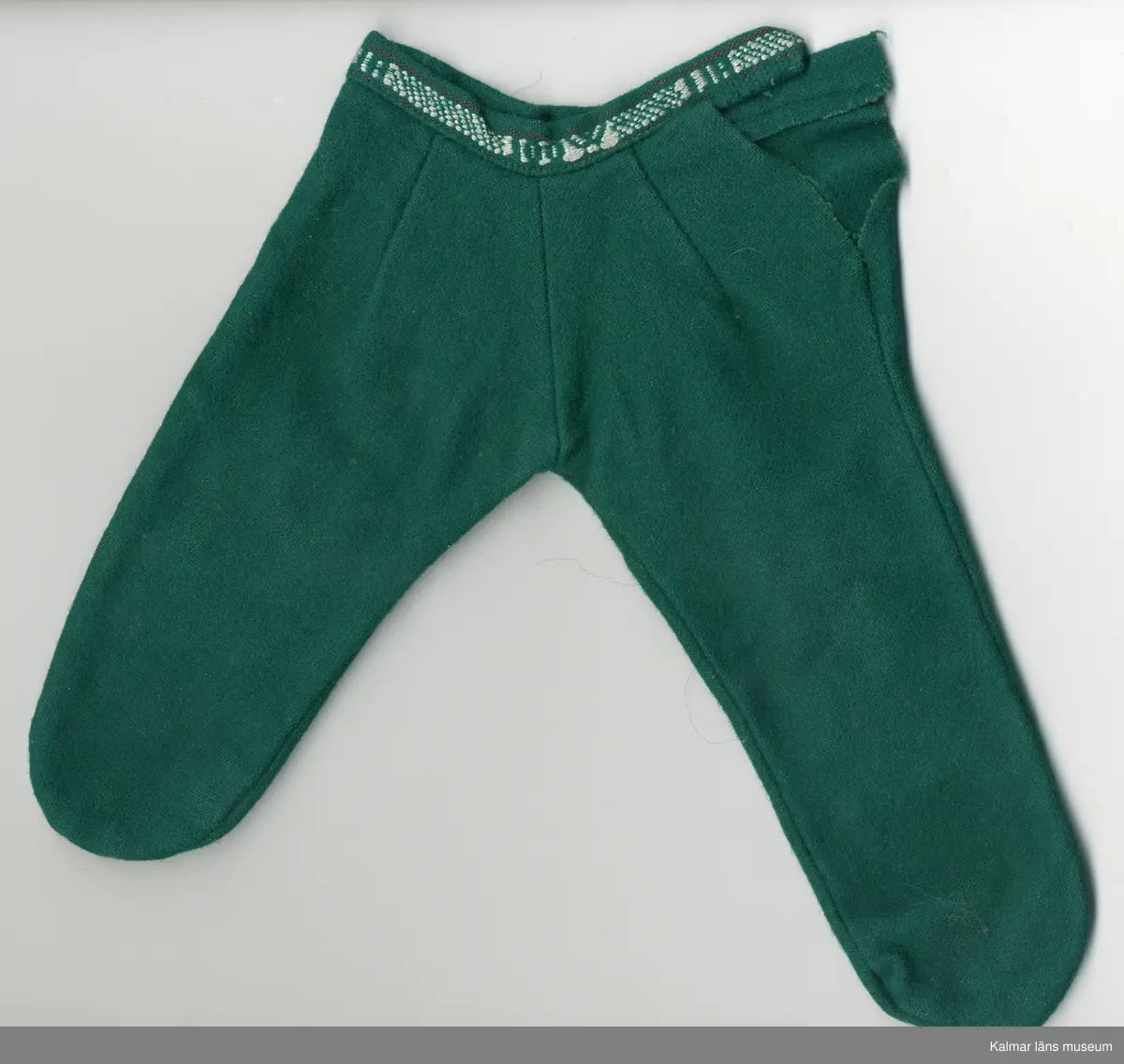 KLM 45519:17:10: 1-3. Dockdräkt, 3 plagg av textil, ylle, grönt. Mönsterbård, invävd, vit och grön med text: ALL WOOL. :1 Dockjacka, rundad halsurringning. dragkedja, framtill, vit. Mönsterbård, två stycken, framtill, längs dragkedja och en bak på jacka, nere vid fållen. :2 Dockbyxa, "sparkbyxa", byxa, hopsydd med rundning vid tår, vänster ben längre. Midja, linning med mönsterbård, knäpps i midja med hake och hyska, av metall. :3 Dockhuvudbonad, skärmmössa, av grönt ylle. Kulle och kant invändigt, förstärkt med Vlieseline, vitt. Mössa, kant, dekorerad med mönsterbård.