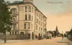 Linköping i ett kolorerat skimmer. Vy utmed S:t Larsgatan från gatans möte med Vasavägen. Det vackra hörnhuset stod klart 1905 och var beställt av Linköpings Litografiska AB. Namnmärket LLAB kan skönjas i fasaden. Längre bort utmed S:t Larsgatan anas S:t Larskyrkan med det uttryck kyrkan fick efter 1906-07 års restaurering. Innanför planket -närmast till höger i bilden- kom Frimurarehotellet att invigas 1912. Sammantaget lämnar dessa nedslag en datering av brevkortets motiv till omkring 1910.