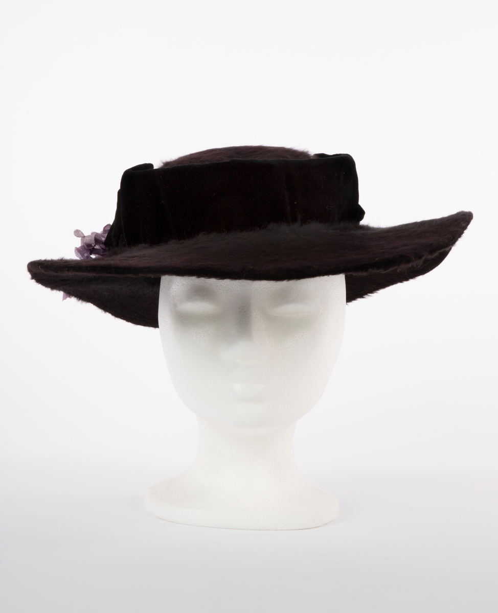 Flat hatt, med lav pull og bred brem. Sydd av sort, glansfull og langhåret hårfilt. Den flate pullen er understøttet av et fullstendig stivt nettstoff og omkranset av et vertikalt "bånd" av sort speilfløyel, avstivet med ståltråd i overkant og trukket over et stivt nettstoff. Hattebåndet har fått innsatt et bredere stykke fløyel over hvert øre, som buer opp i en  spiss, også disse avstivet og formet av en innvendig ståltråd. Den brede bremmen er påsydd og ombrettet i ytterkant, slik at den lodne filten også sees fra undersiden, der den er festet med overbrett på innsiden av pullens nederkant. For ekstra stivhet, er bremmens ytterkant innvendig avstivet med 2 tettstilte, trådomspunne, svært tynne ståltråder, sydd parallelt med ytterkanten. Bak på bremmen, litt til høyre, hviler en stor bukett fioler av tynt, stivt tekstil. Innvendig trukket med svart fôrsilke, rynket tett sammen i toppen. Både brem og fôr er festet for hånd med svart tråd, mens fiolbuketten er røft festet med blekgrønn tråd av samme farge som stilkene.

Det kan se ut som om garnering med speilfløyel og fiolbukett kan være sekundær.