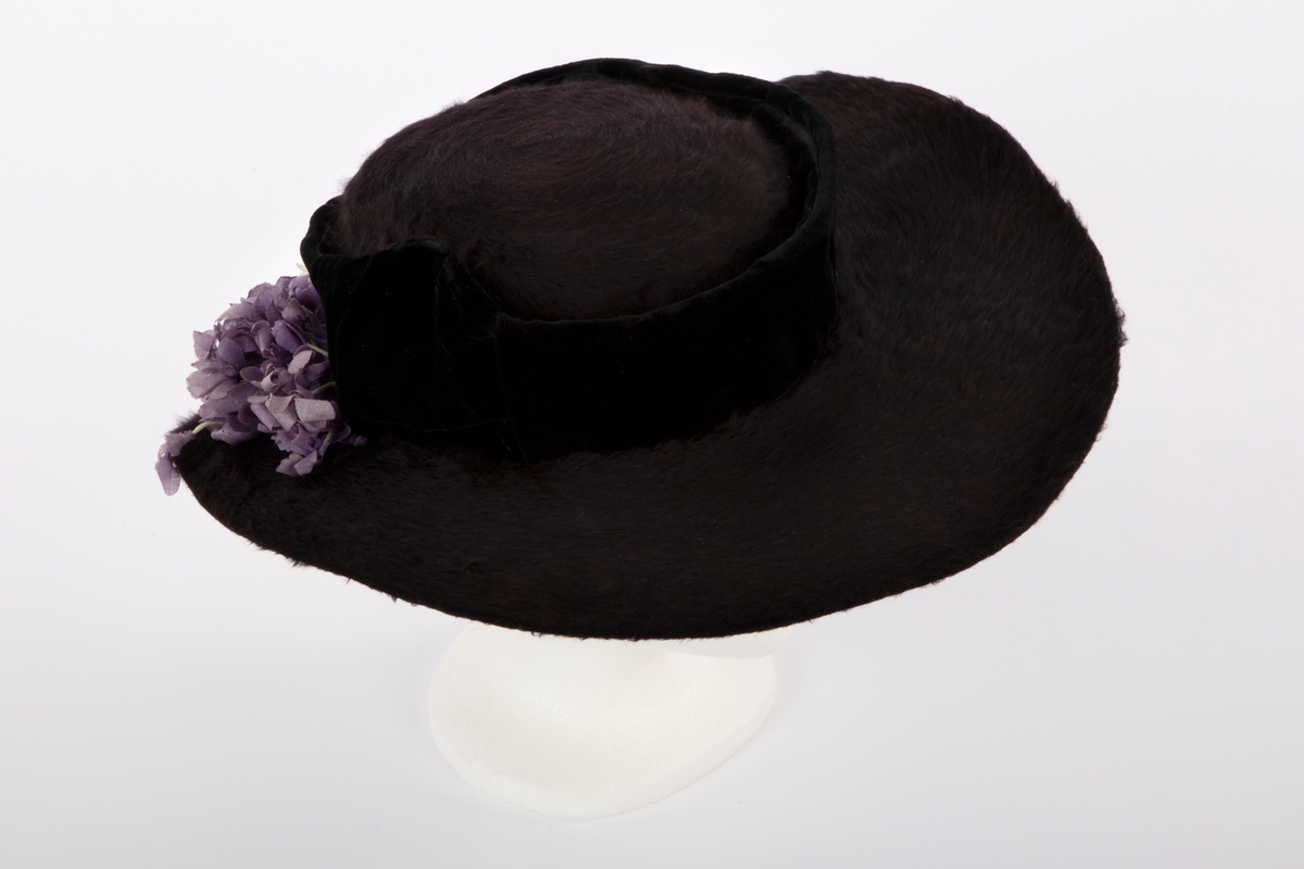 Flat hatt, med lav pull og bred brem. Sydd av sort, glansfull og langhåret hårfilt. Den flate pullen er understøttet av et fullstendig stivt nettstoff og omkranset av et vertikalt "bånd" av sort speilfløyel, avstivet med ståltråd i overkant og trukket over et stivt nettstoff. Hattebåndet har fått innsatt et bredere stykke fløyel over hvert øre, som buer opp i en  spiss, også disse avstivet og formet av en innvendig ståltråd. Den brede bremmen er påsydd og ombrettet i ytterkant, slik at den lodne filten også sees fra undersiden, der den er festet med overbrett på innsiden av pullens nederkant. For ekstra stivhet, er bremmens ytterkant innvendig avstivet med 2 tettstilte, trådomspunne, svært tynne ståltråder, sydd parallelt med ytterkanten. Bak på bremmen, litt til høyre, hviler en stor bukett fioler av tynt, stivt tekstil. Innvendig trukket med svart fôrsilke, rynket tett sammen i toppen. Både brem og fôr er festet for hånd med svart tråd, mens fiolbuketten er røft festet med blekgrønn tråd av samme farge som stilkene.

Det kan se ut som om garnering med speilfløyel og fiolbukett kan være sekundær.