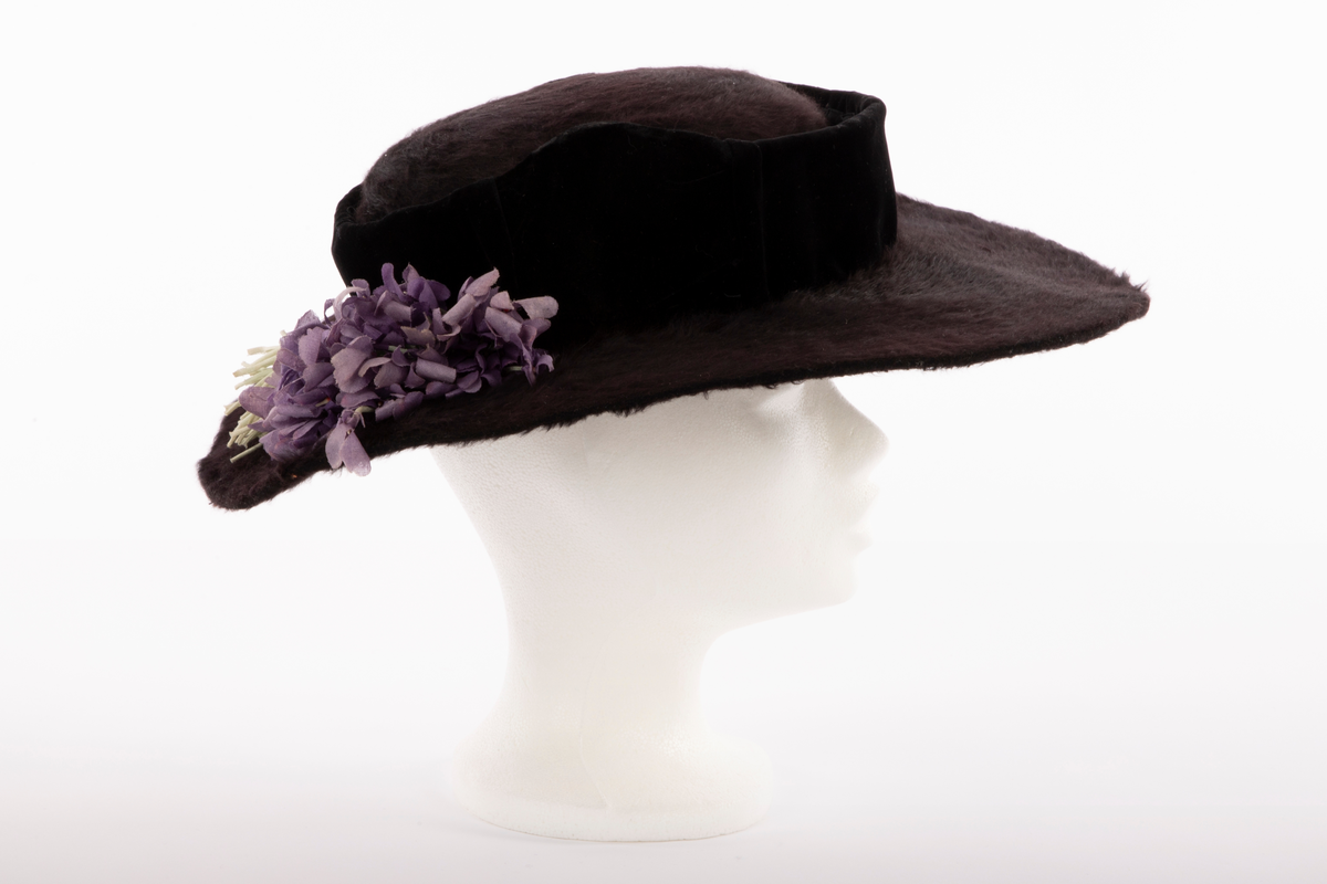 Flat hatt, med lav pull og bred brem. Sydd av sort, glansfull og langhåret hårfilt. Den flate pullen er understøttet av et fullstendig stivt nettstoff og omkranset av et vertikalt "bånd" av sort speilfløyel, avstivet med ståltråd i overkant og trukket over et stivt nettstoff. Hattebåndet har fått innsatt et bredere stykke fløyel over hvert øre, som buer opp i en  spiss, også disse avstivet og formet av en innvendig ståltråd. Den brede bremmen er påsydd og ombrettet i ytterkant, slik at den lodne filten også sees fra undersiden, der den er festet med overbrett på innsiden av pullens nederkant. For ekstra stivhet, er bremmens ytterkant innvendig avstivet med 2 tettstilte, trådomspunne, svært tynne ståltråder, sydd parallelt med ytterkanten. Bak på bremmen, litt til høyre, hviler en stor bukett fioler av tynt, stivt tekstil. Innvendig trukket med svart fôrsilke, rynket tett sammen i toppen. Både brem og fôr er festet for hånd med svart tråd, mens fiolbuketten er røft festet med blekgrønn tråd av samme farge som stilkene.

Det kan se ut som om garnering med speilfløyel og fiolbukett kan være sekundær.