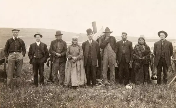 Historisk bilde av ni mennesker, syv menn og to kvinner stilt opp på rekke. Mennene har hatt. Kvinnene og tre av mennene ser ut til å være urbefolkning. Bak gruppa stikker et ovnsrør opp fra et telt. 