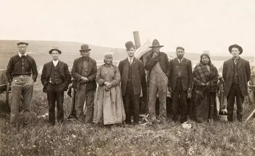 Historisk bilde av ni mennesker, syv menn og to kvinner stilt opp på rekke. Mennene har hatt. Kvinnene og tre av mennene ser ut til å være urbefolkning. Bak gruppa stikker et ovnsrør opp fra et telt. 