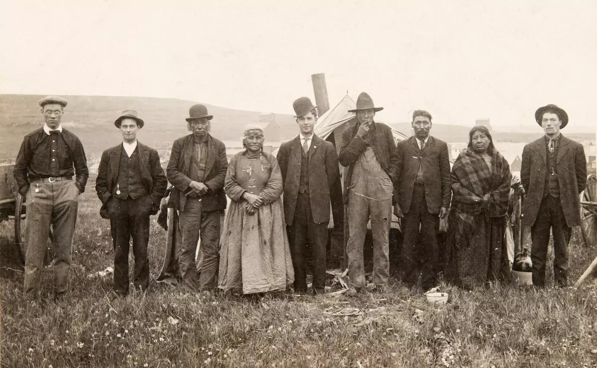 Historisk bilde av ni mennesker, syv menn og to kvinner stilt opp på rekke. Mennene har hatt. Kvinnene og tre av mennene ser ut til å være urbefolkning. Bak gruppa stikker et ovnsrør opp fra et telt. 