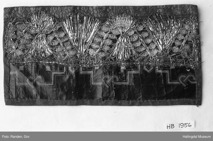 Voksduk med mønster i gull og grått. Påsatt bred sølvknipling. Øverst blomsterete bånd. Foret med sort tøy. Tråklesting i sort og hvitt.