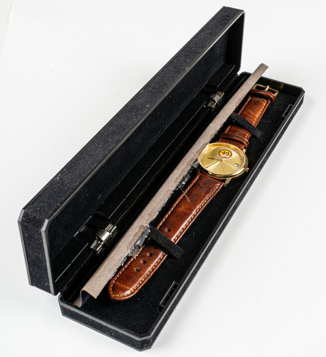 Armbandsklocka i svart etui i sammet med guldtryck. Klocka med guldfärgad urtavla, datumvisare, jubileumsmärke samt en rosa Ahlgrens bil. Brunt läderarmband.