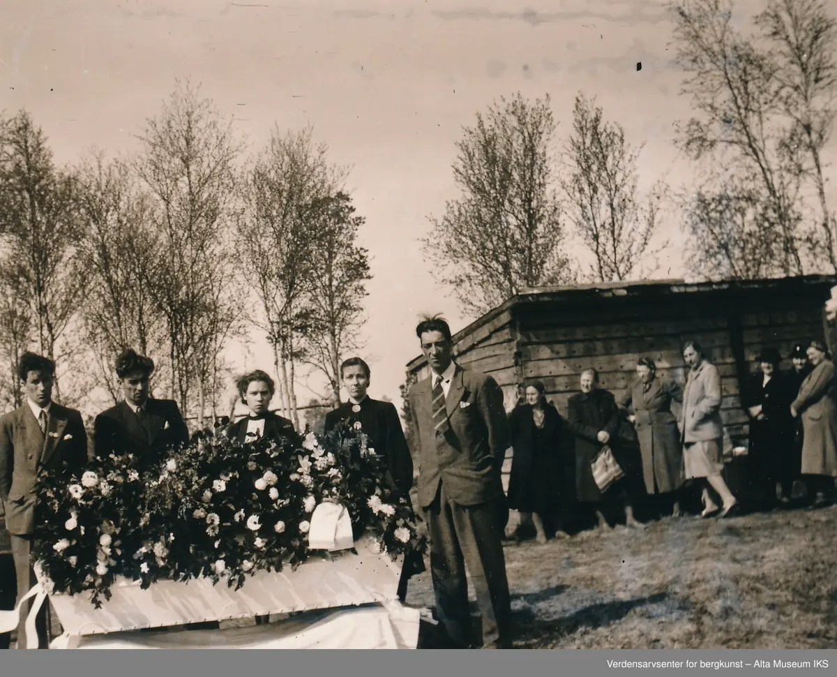 Begravelse med kiste dekket av blomster og familie samlet rundt.