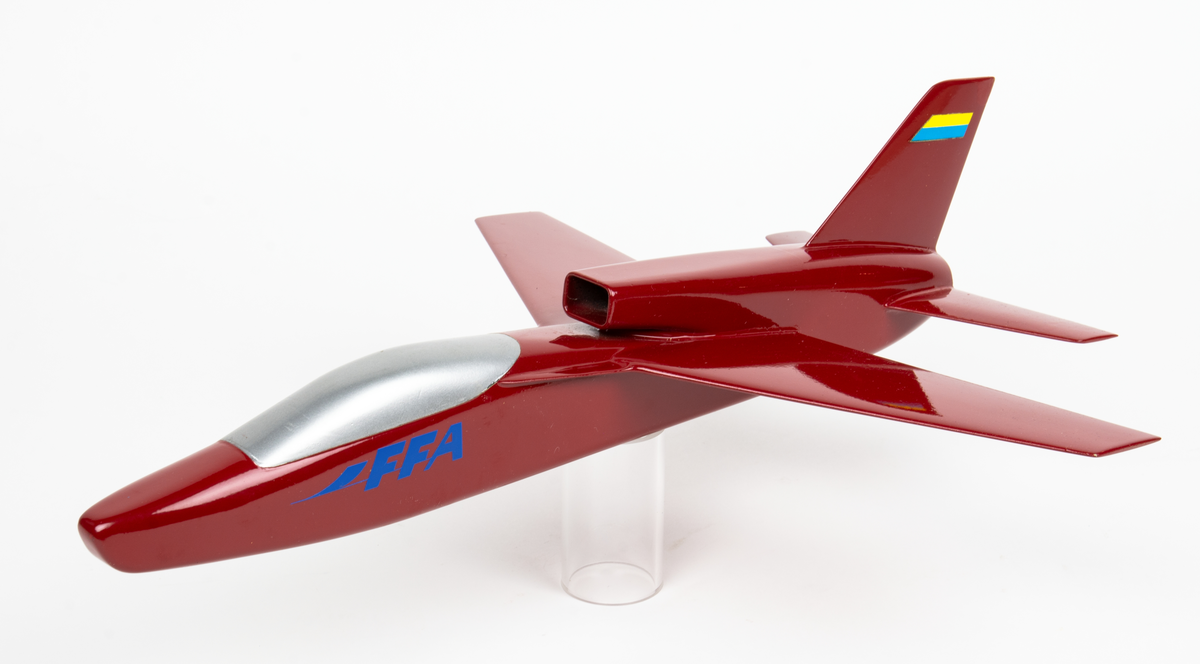 Flygplansmodell av B3LA, Saab 38,  med ryggintag. Röd trämodell med texten FFA på sidan samt en gul och blå märkning på fenan bak.
Modellen förvaras i en specialtillverkad trälåda.