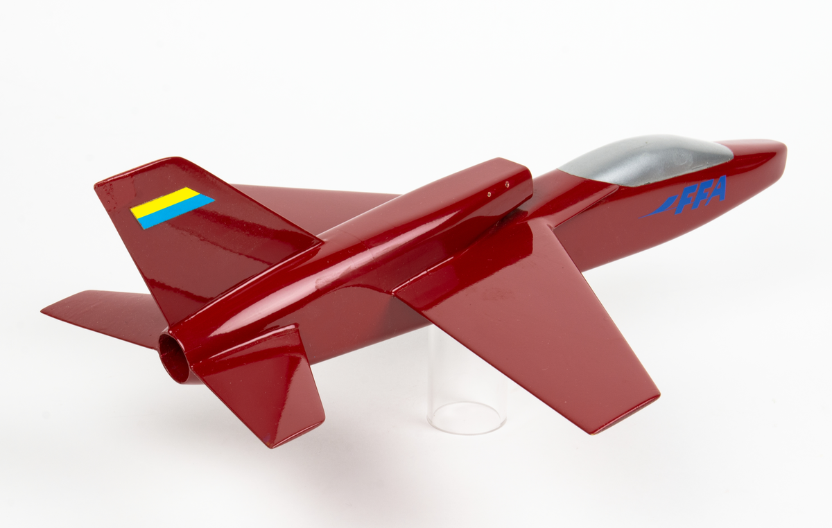 Flygplansmodell av B3LA, Saab 38,  med ryggintag. Röd trämodell med texten FFA på sidan samt en gul och blå märkning på fenan bak.
Modellen förvaras i en specialtillverkad trälåda.