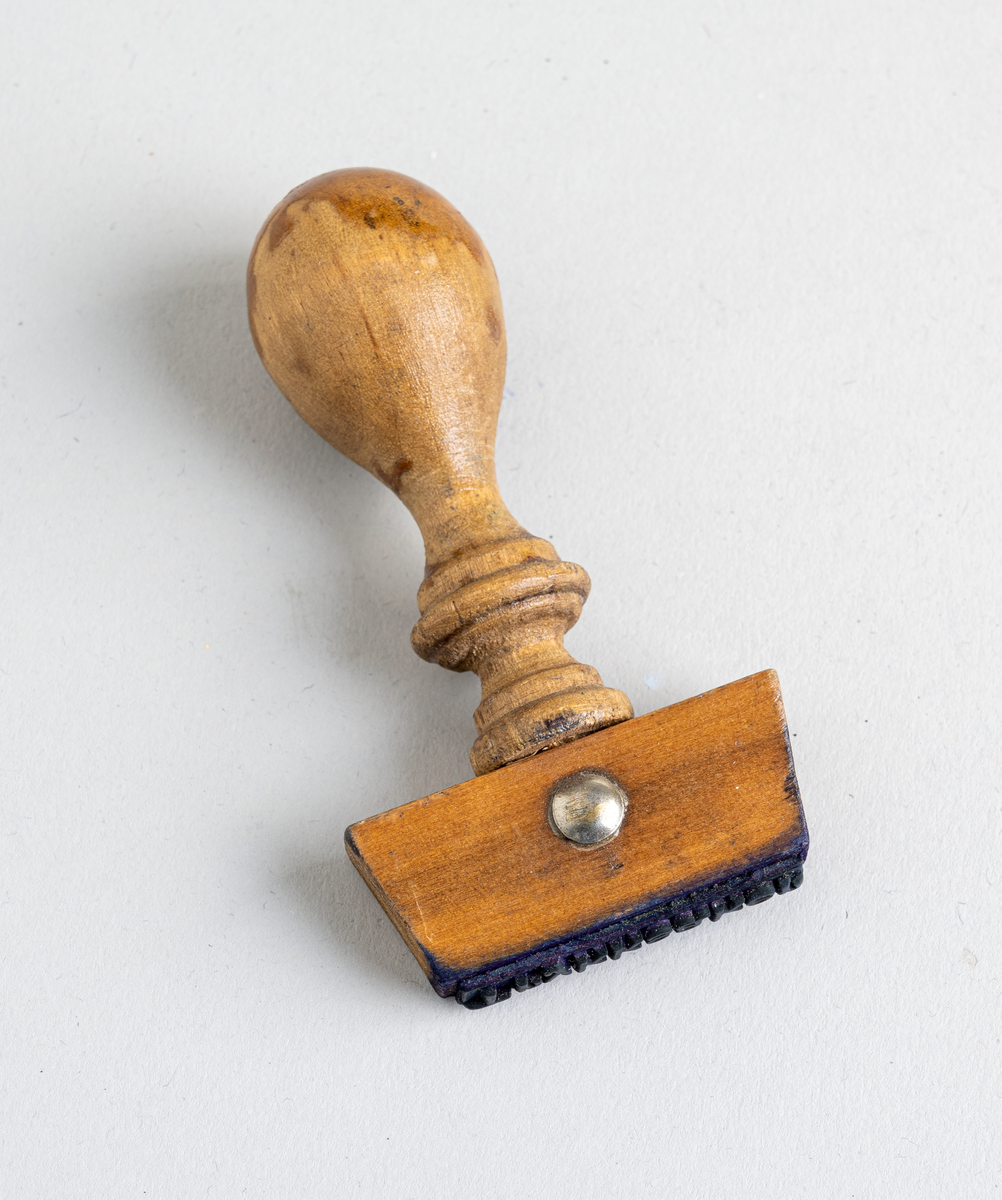 Et stempel fra Nordenfjeldske Bjergingskompani. Stempelet er en trapesformet trekloss. Et dråpeformet håndtak er festet på midten. Håndtaket har dreid dekor. Alt treverk er lakkert. Stempeldelen er av gummi.
Tekst på stempelet: Trykksaker