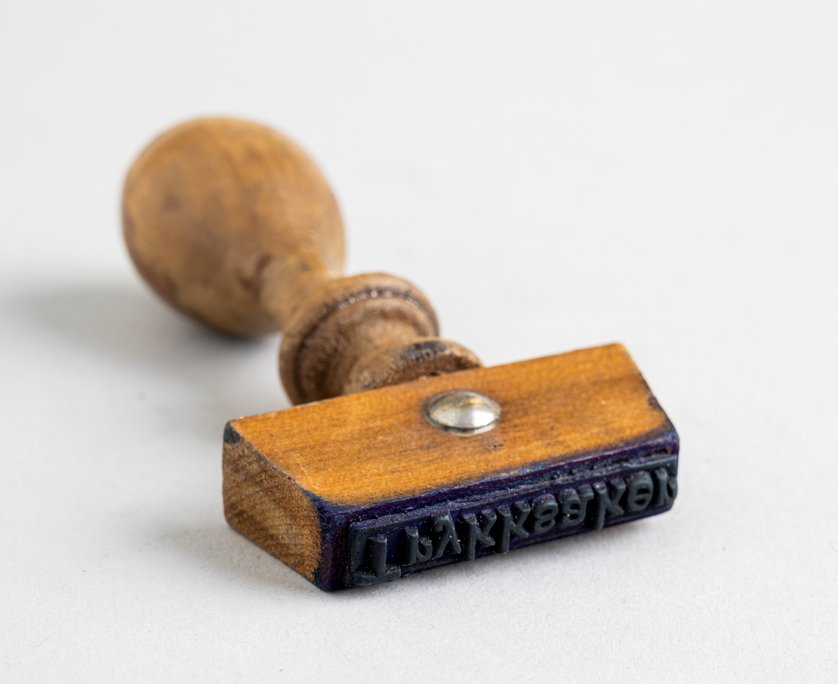 Et stempel fra Nordenfjeldske Bjergingskompani. Stempelet er en trapesformet trekloss. Et dråpeformet håndtak er festet på midten. Håndtaket har dreid dekor. Alt treverk er lakkert. Stempeldelen er av gummi.
Tekst på stempelet: Trykksaker