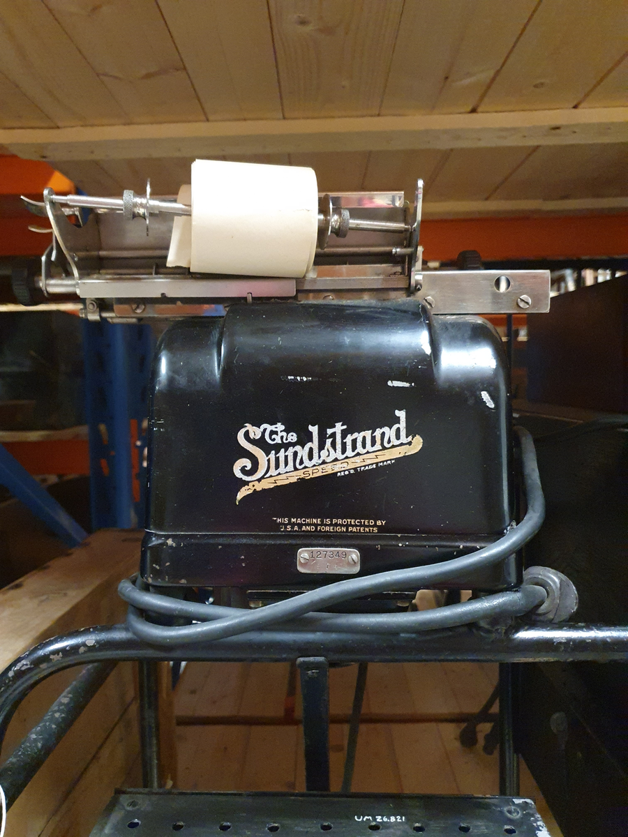 Räknemaskin på rullbord. Elektrisk motor och elsladd. Svartmålad plåt. Bordsbricka på bordet, kan fällas. Räknemaskinen är 20 cm bred, 15 cm hög, 37 cm djup. Handdriven vals med isatt pappersrulle. Knappsats med röda och svarta knappar. Etikett "The Sundstrand" REG´D TRADE MARK TEN, THOU, MILL, COLUMNS LISTING 0123456789, 9 column totals.