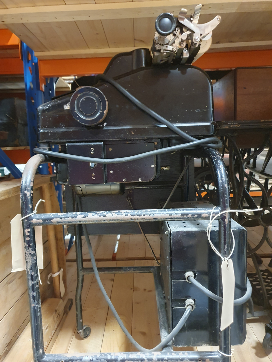 Räknemaskin på rullbord. Elektrisk motor och elsladd. Svartmålad plåt. Bordsbricka på bordet, kan fällas. Räknemaskinen är 20 cm bred, 15 cm hög, 37 cm djup. Handdriven vals med isatt pappersrulle. Knappsats med röda och svarta knappar. Etikett "The Sundstrand" REG´D TRADE MARK TEN, THOU, MILL, COLUMNS LISTING 0123456789, 9 column totals.