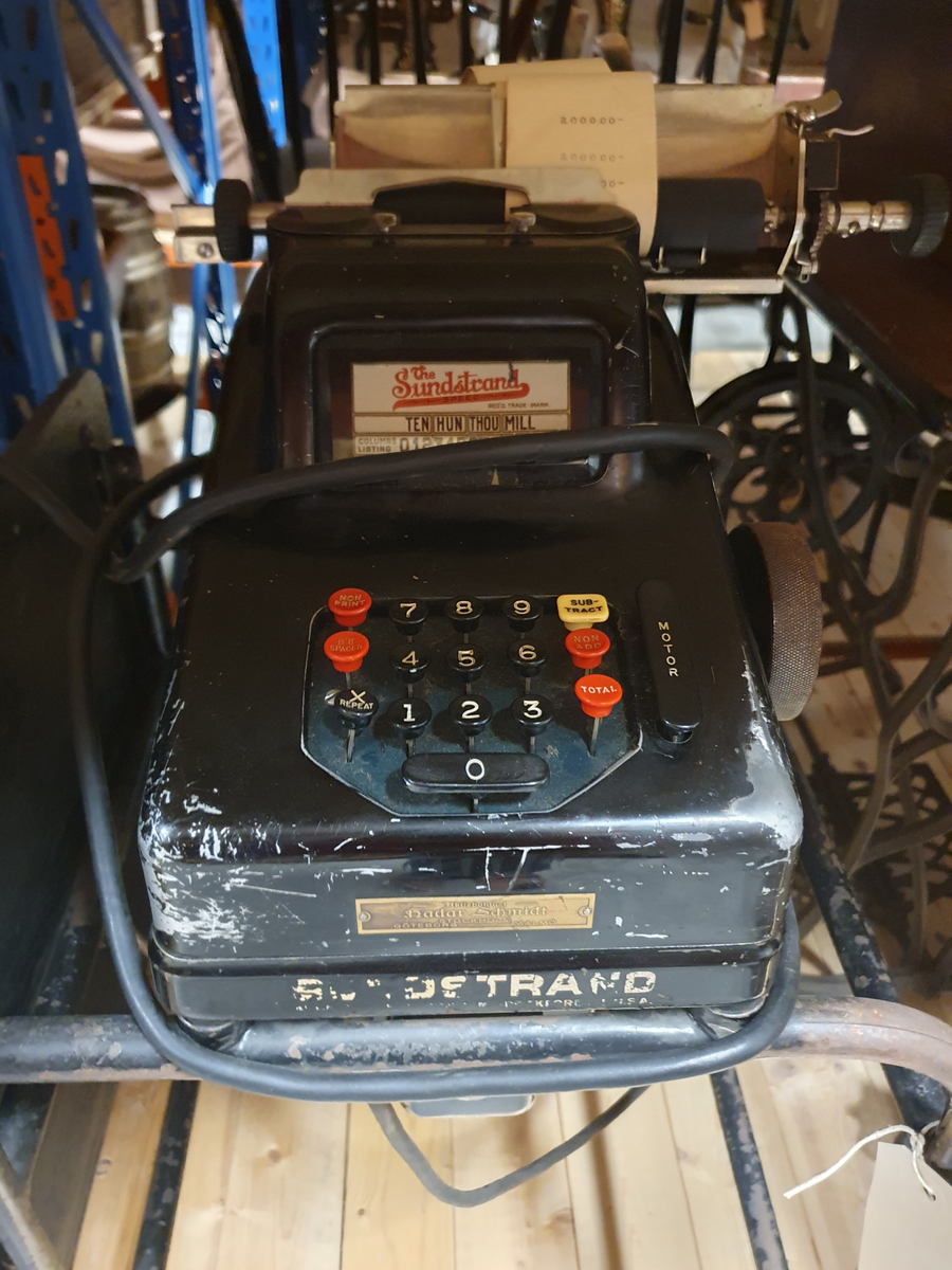 Räknemaskin på rullbord. Elektrisk motor och elsladd. Svartmålad plåt. Bordsbricka på bordet, kan fällas. Räknemaskinen är 20 cm bred, 15 cm hög, 37 cm djup. Handdriven vals med isatt pappersrulle. Knappsats med röda och svarta knappar. Etikett "The Sundstrand" REG´D TRADE MARK TEN, THOU, MILL, COLUMNS LISTING 0123456789, 9 column totals.