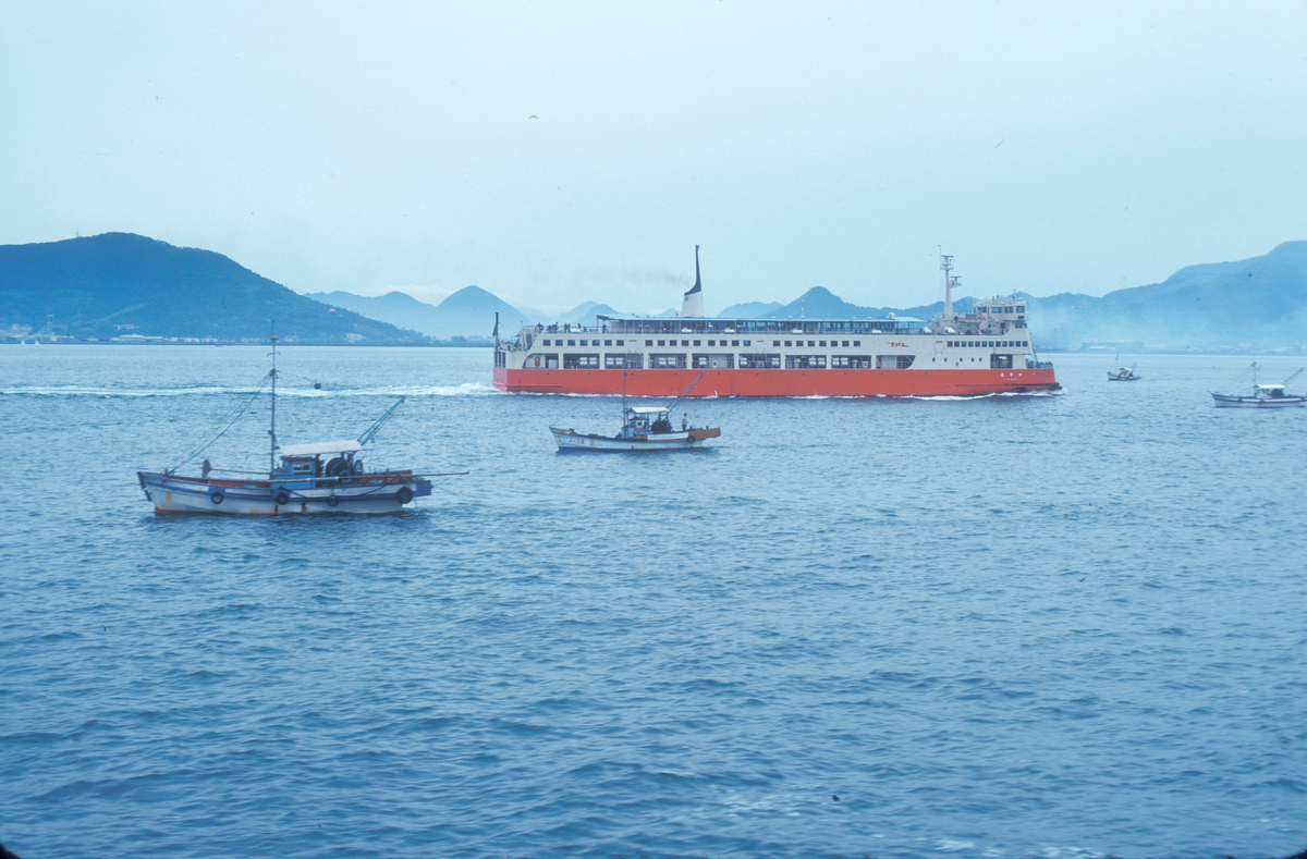 Motiv fra Japantur : Fiskebåter, og en passasjerbåt(?) i fart, med kystlandskap i bakgrunnen