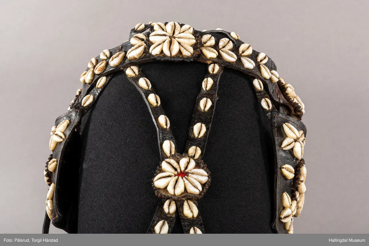 Hodelag til hest med munnbitt og tømmer. Hodelaget er dekoret med kauriskjell og rød ull. Skylapper, trapesformet dekorert midt på med lærrosett og hvite kauriskjell. Kort tømme, ender i kute av lærfletting og røde ullfrynser, frynsene er slitt bort. Munnbitt av jern. Stassele.