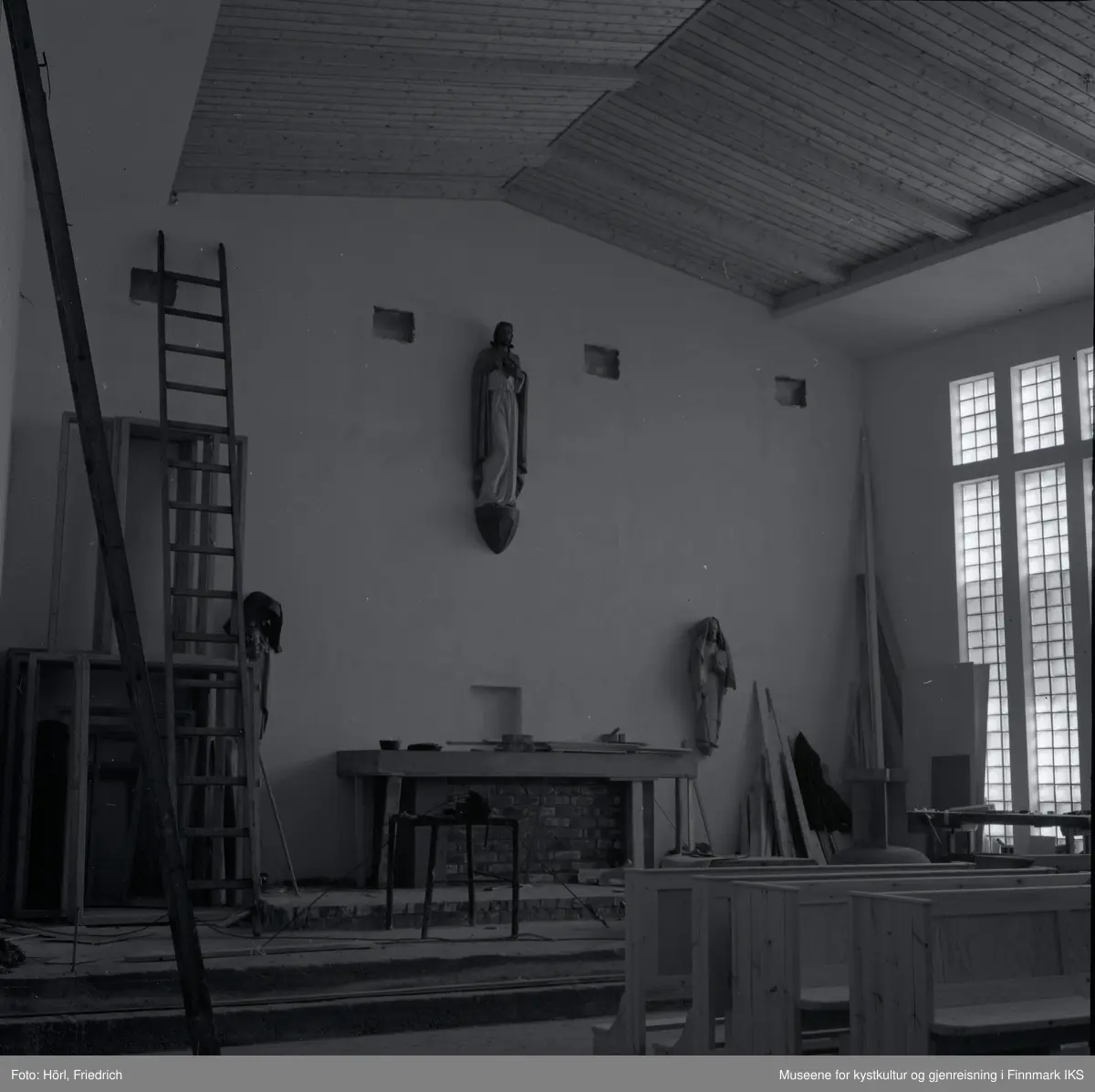 Interiøret til den katolske kirka St. Mikael i Hammerfest bygges i 1958. I motsetning til senere, er det montert en skulptur av Jesus Kristus ovenfor alteret. Senere ble det plassert et krusifiks der. Mosaikken på fronten av alteret som ble utført av Karl Manninger, er ikke kommet på plass enda. Til venstre for alteret er det montert en skulptur av Maria med barnet, til høyre vises Josef. Disse to skulpturene som er skjært av tre er tildekket med tepper. Foran alteret er det satt opp en bordsag. Til venstre i bilde står en lang stige og noen rammer lent til veggen, til høyre ved vinduene ser man en høvelbenk og mer byggemateriale. I forgrunnen til høyre står det kirkebenker.