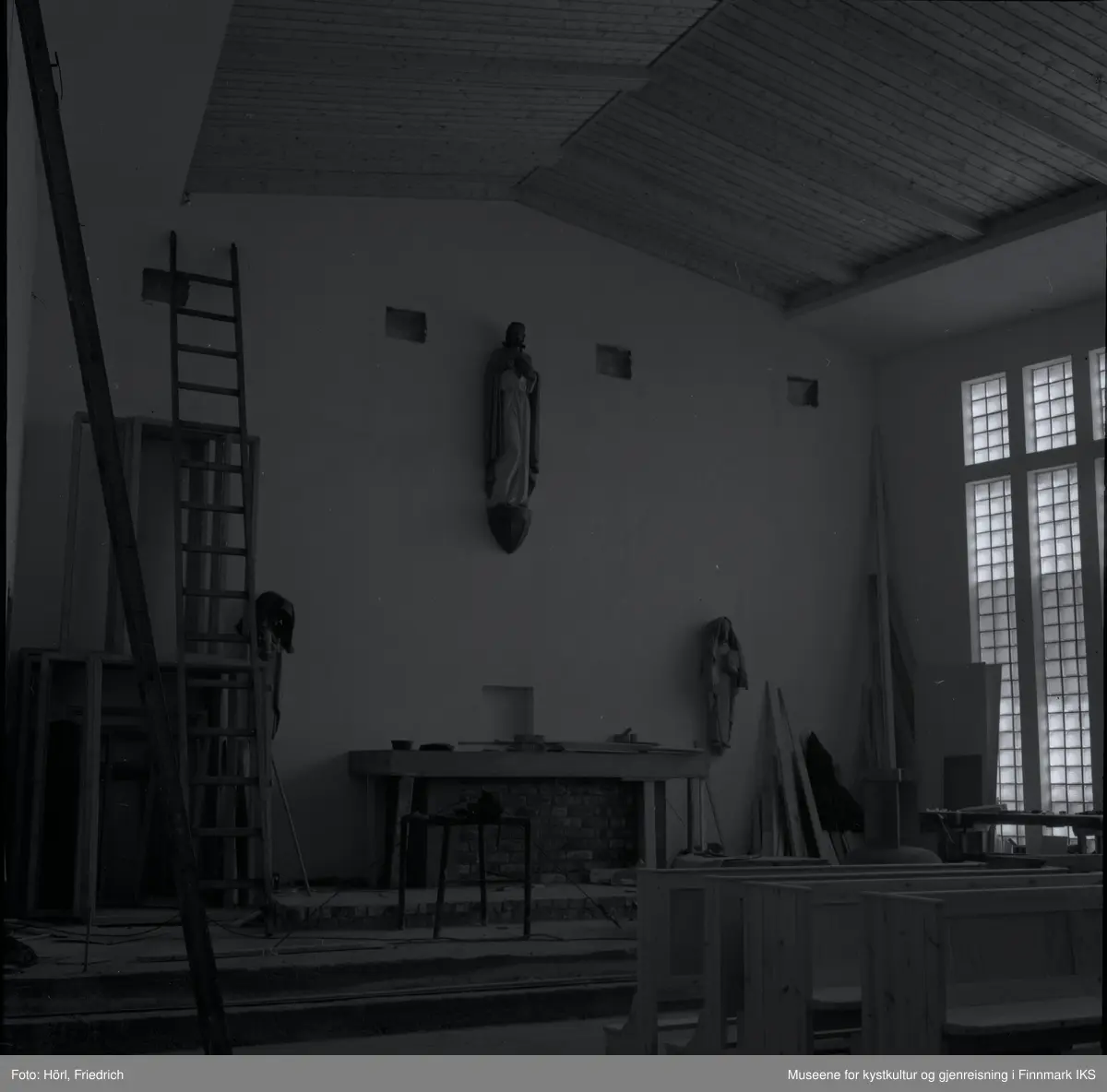 Interiøret til den katolske kirka St. Mikael i Hammerfest bygges i 1958. I motsetning til senere, er det montert en skulptur av Jesus Kristus ovenfor alteret. Senere ble det plassert et krusifiks der. Mosaikken på fronten av alteret som ble utført av Karl Manninger, er ikke kommet på plass enda. Til venstre for alteret er det montert en skulptur av Maria med barnet, til høyre vises Josef. Disse to skulpturene som er skjært av tre er tildekket med tepper. Foran alteret er det satt opp en bordsag. Til venstre i bilde står en lang stige og noen rammer lent til veggen, til høyre ved vinduene ser man en høvelbenk og mer byggemateriale. I forgrunnen til høyre står det kirkebenker.